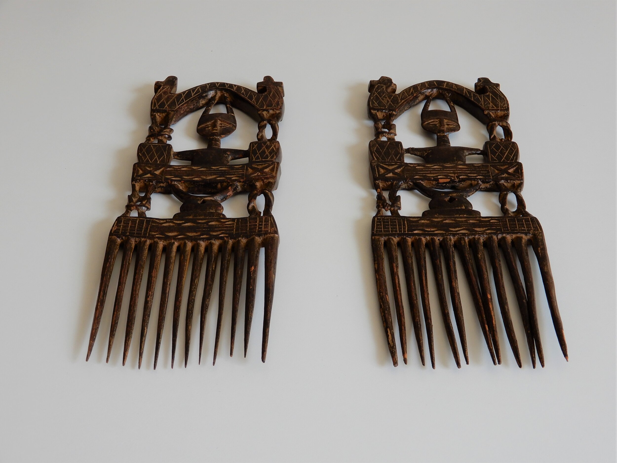  A pair of dark wooden combs. Each comb is made up of three vertical sections: the top section shows an abstract humanoid figure under an arch. The second section has a stool which holds up the floor which the humanoid figure is on. The third section