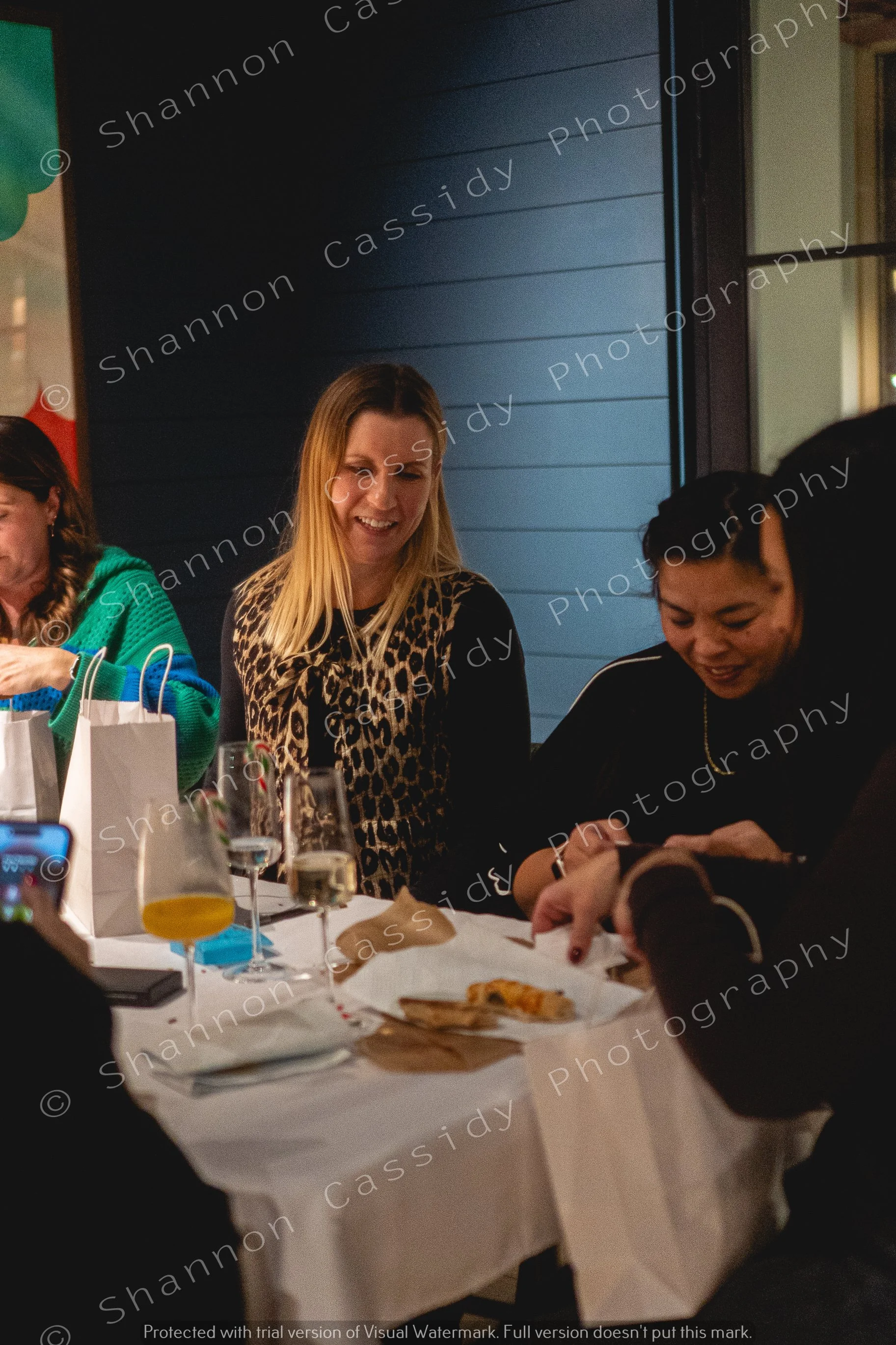 People sitting at a dinner table with glasses of drinks and food, smiling and socializing in a dimly lit room.