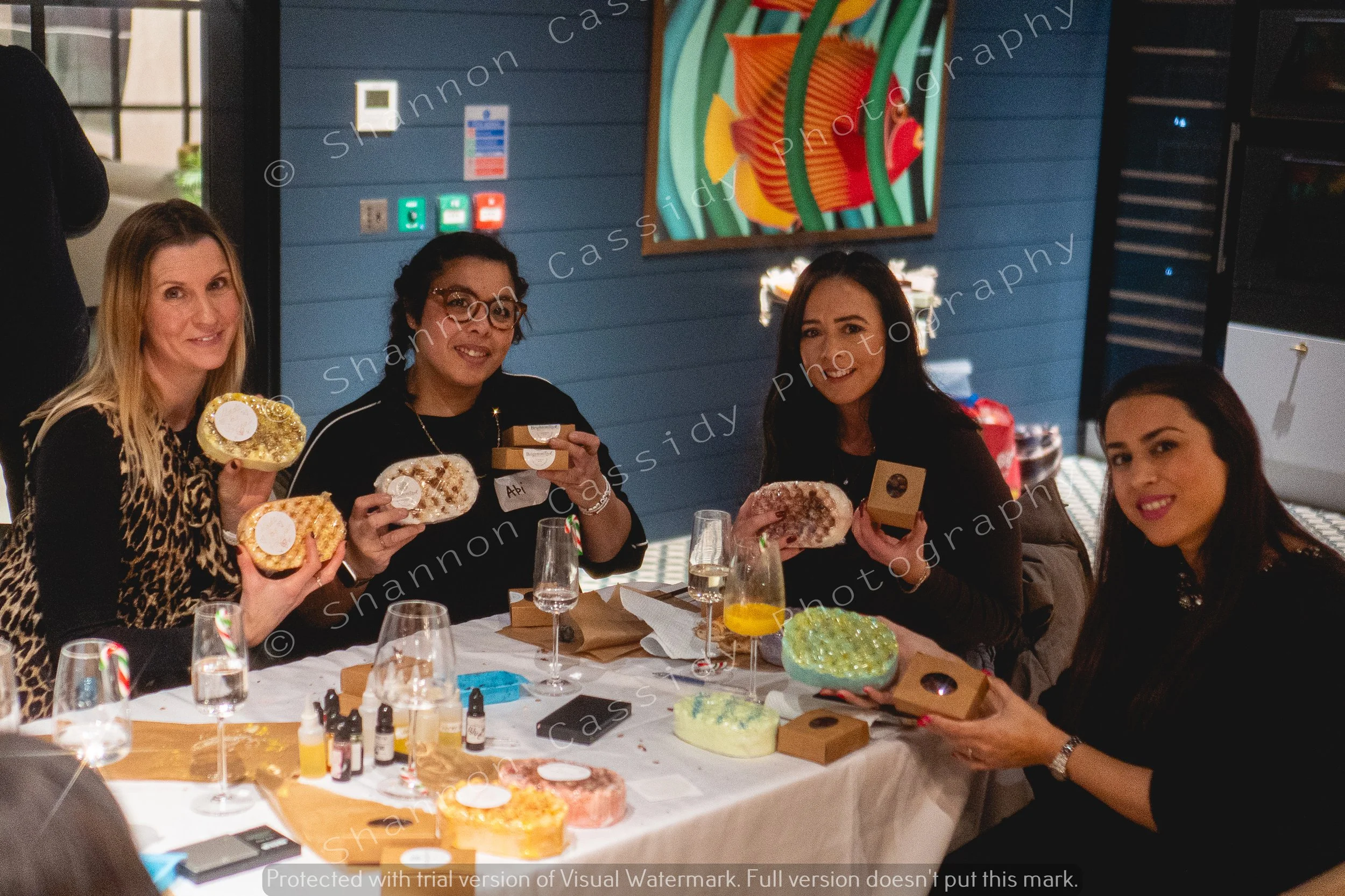 Four women sitting around a table, holding decorated cakes with colorful frosting and edible decorations, with various boxes and drinks on the table, in a decorated indoor setting.