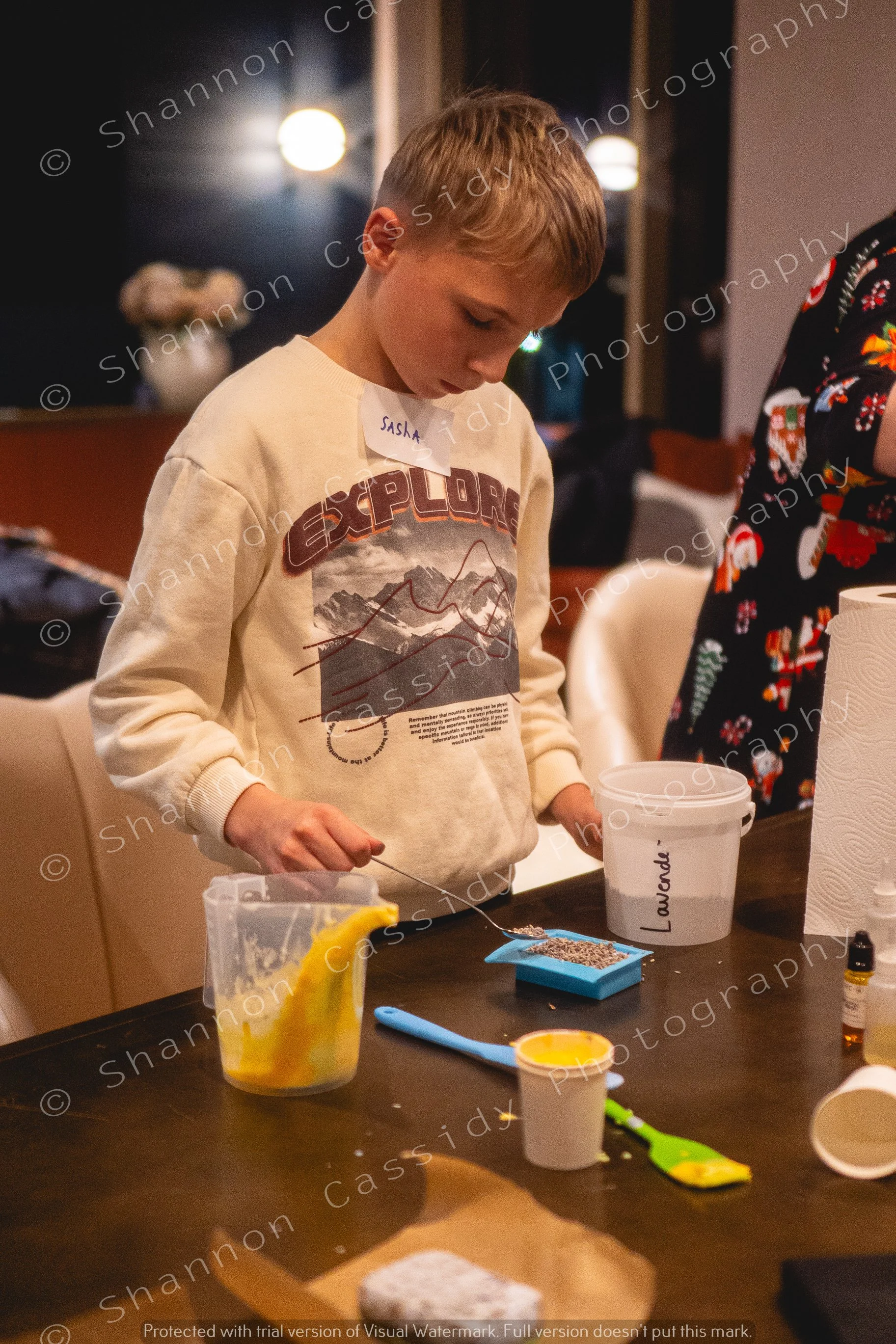 A young boy with a name tag that reads Sasha stands at a table with baking ingredients and tools, focusing on a task during a cooking or baking activity in a warm, indoor setting.
