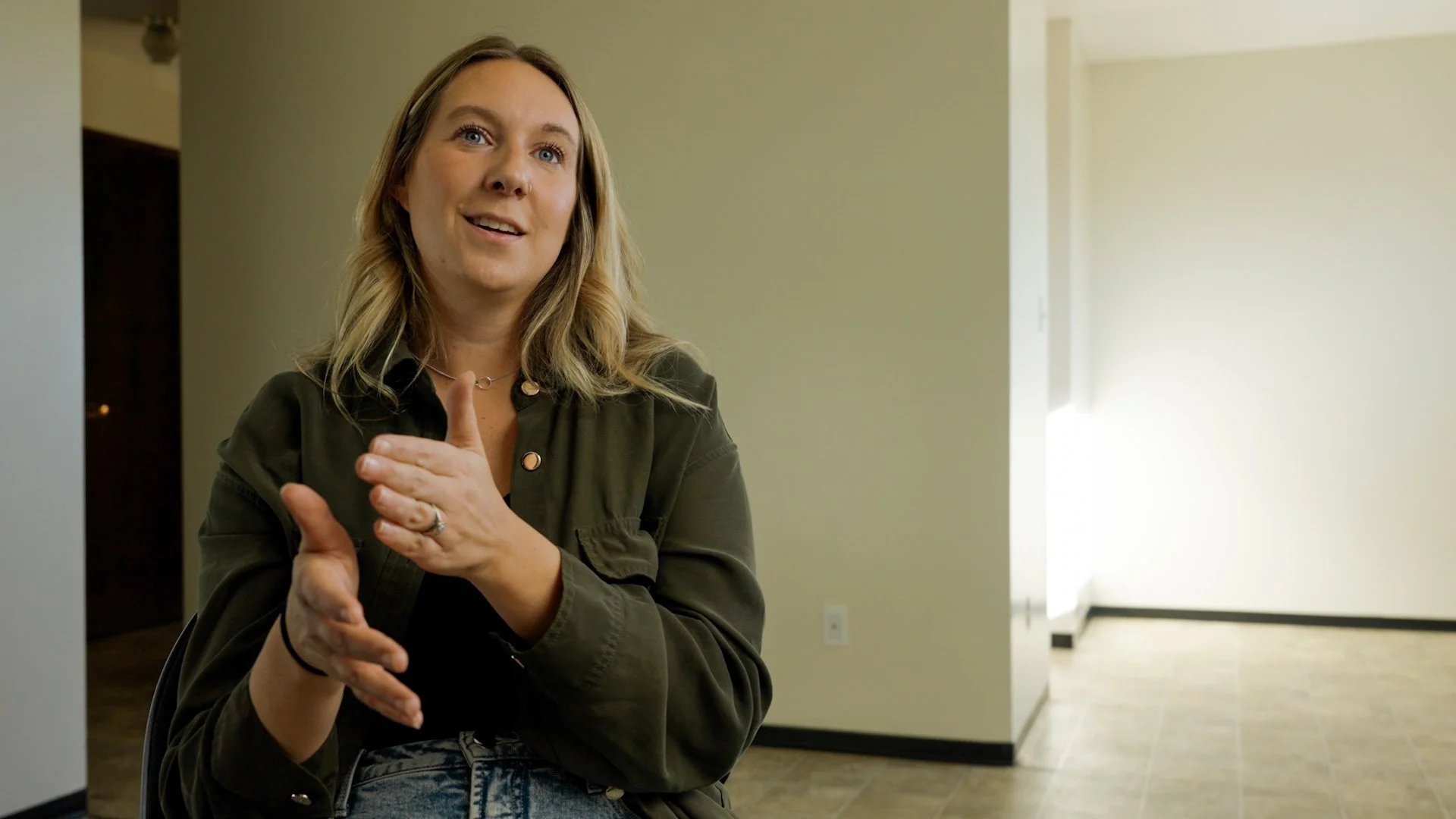 An older woman with glasses and silver hair sitting in a building with modern decor, a large window with a view outside, and a white wall with text in the background.