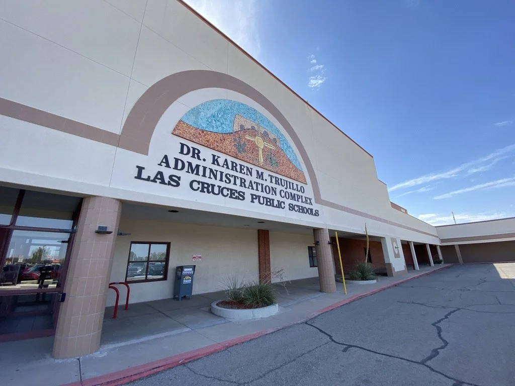 Front of Dr. Karen M. Trujillo Administration Complex at Las Cruces Public Schools, with a mosaic sign on the building, clear blue sky, and a parking lot in the foreground.