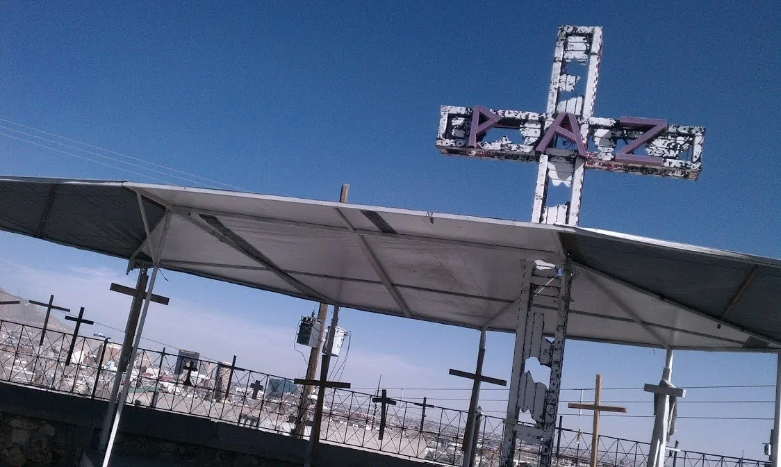 Metal cross with the word 'GRAZ' in purple letters, under a large metal canopy, with multiple smaller crosses and electrical poles in the background, against a clear blue sky.