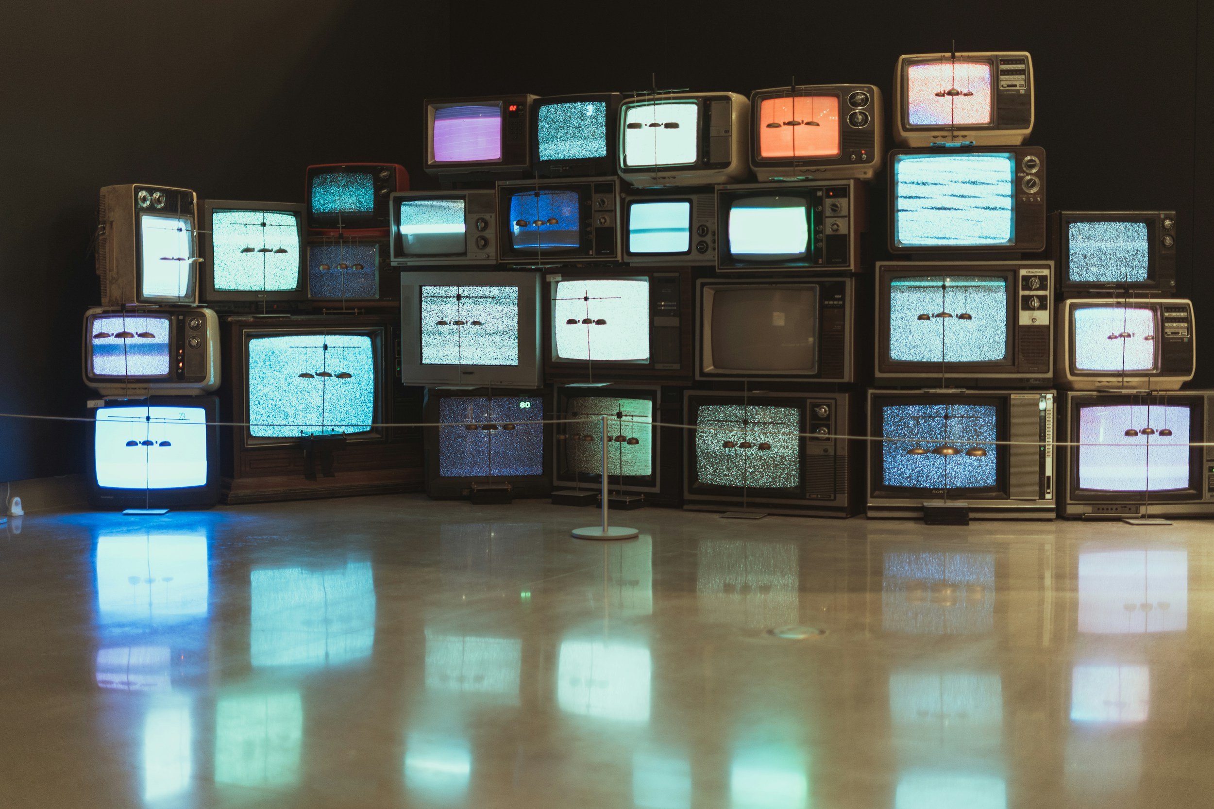 A collection of vintage televisions stacked against a dark background, some displaying static and others turned off, with a reflective floor creating mirror images of the screens.
