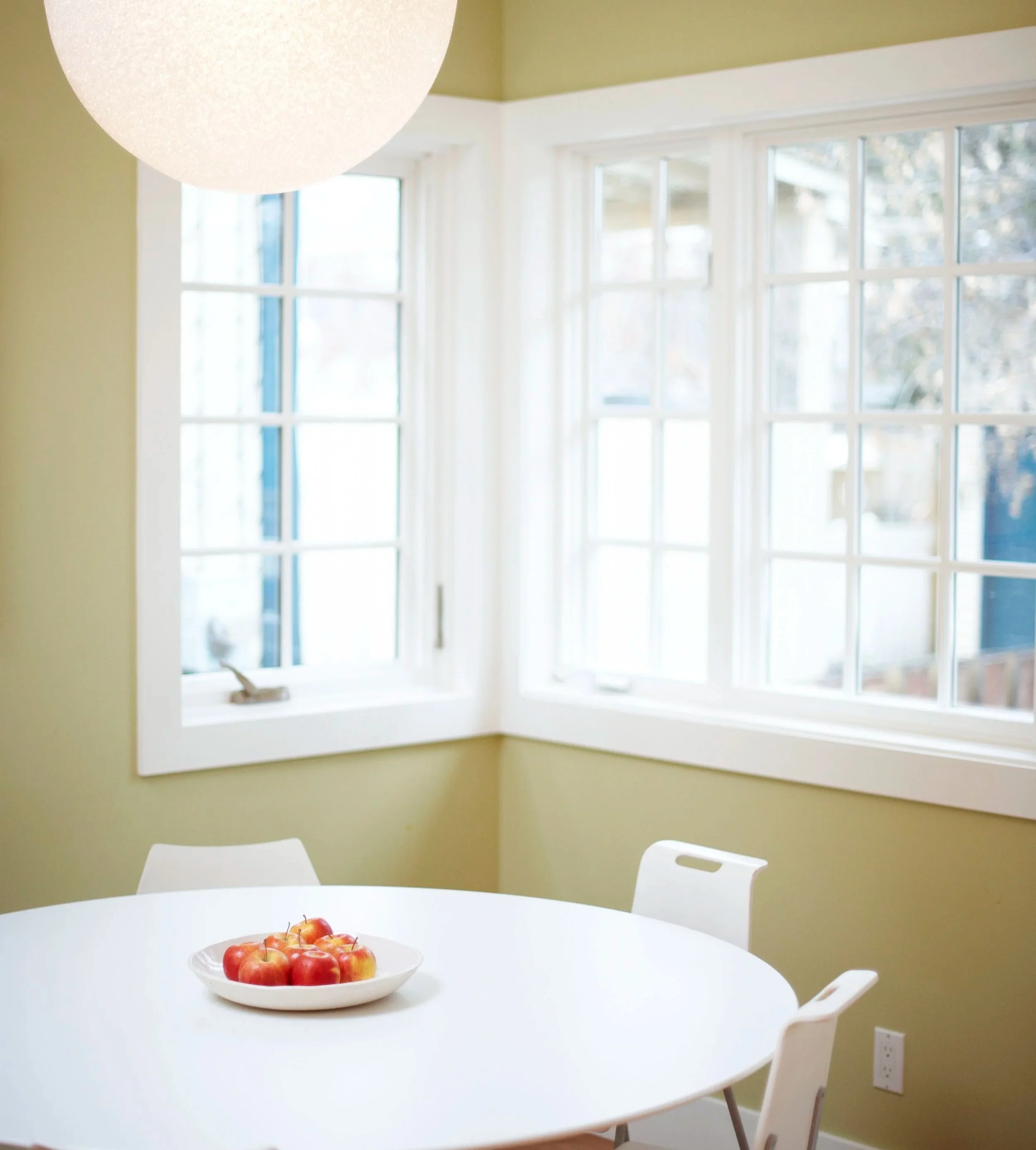 Dining area with a white round table, a plate of red apples, white chairs, large corner windows, and a hanging ceiling light.
