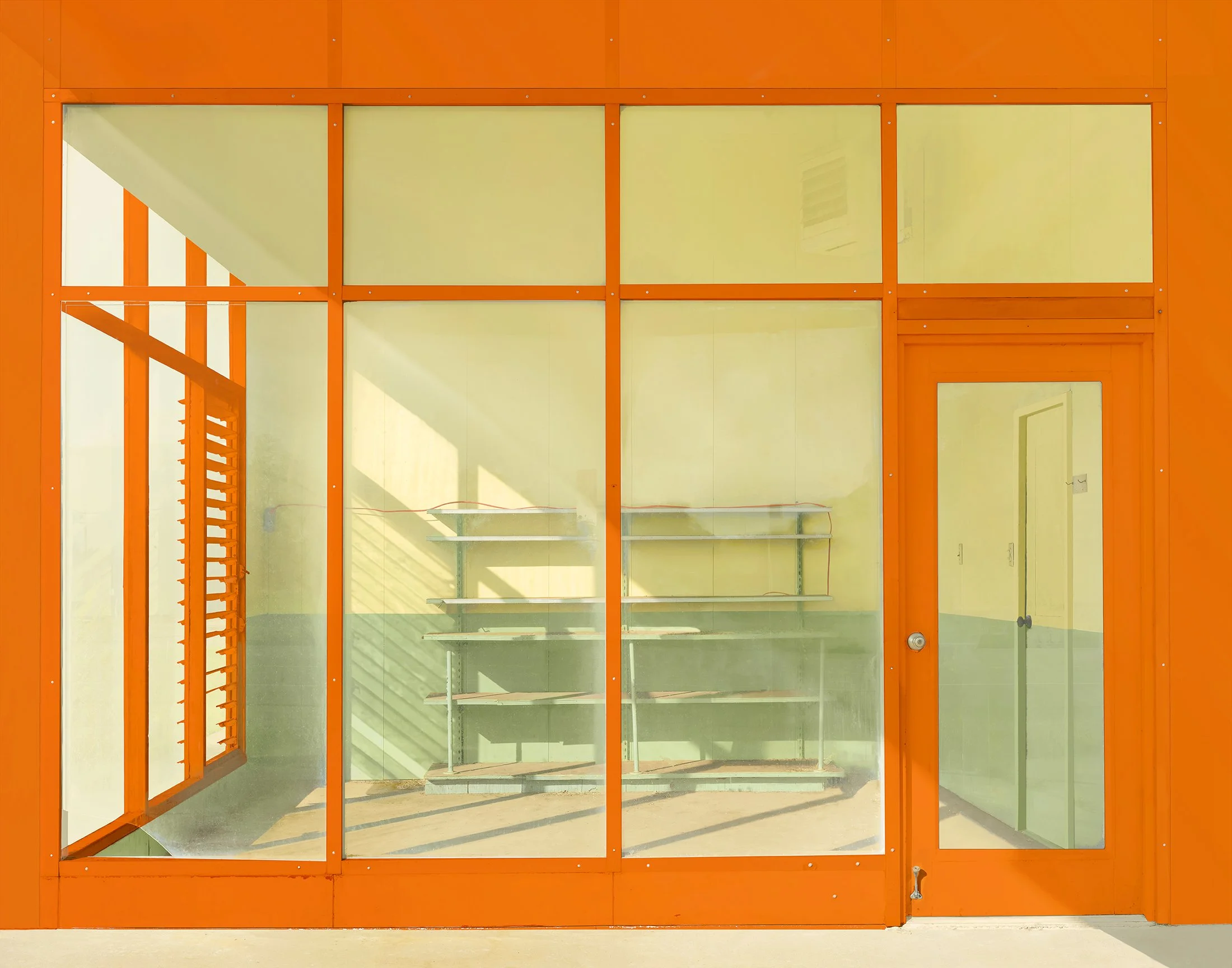 Photo of a bright orange-framed glass door and window with a view of an unfinished interior space with green walls, metal shelving, and electrical wiring.