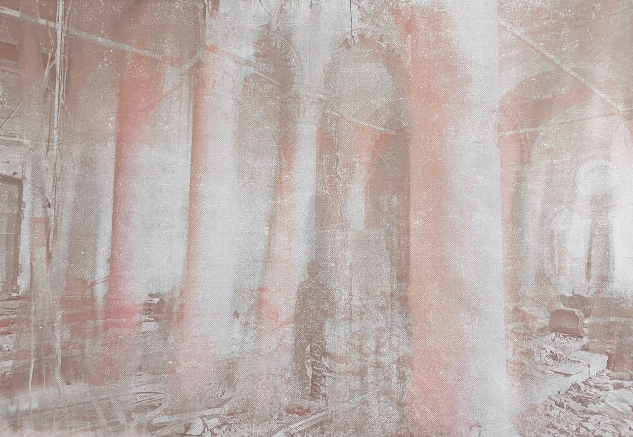 A smartphone photo taken inside a building with large columns and damaged walls, showing debris on the floor and a person standing in the background.