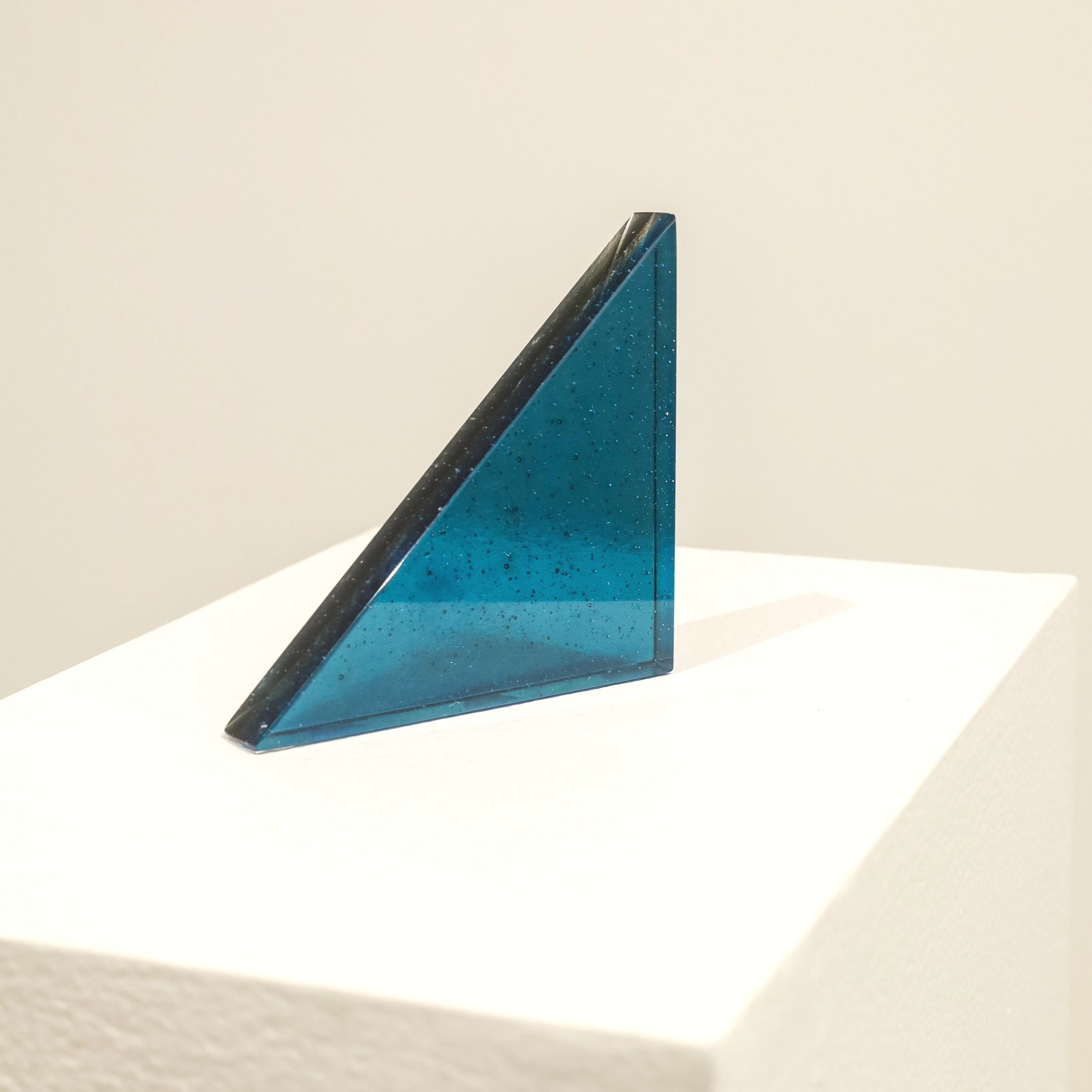 A blue triangular glass sculpture displayed on a white pedestal in a gallery.
