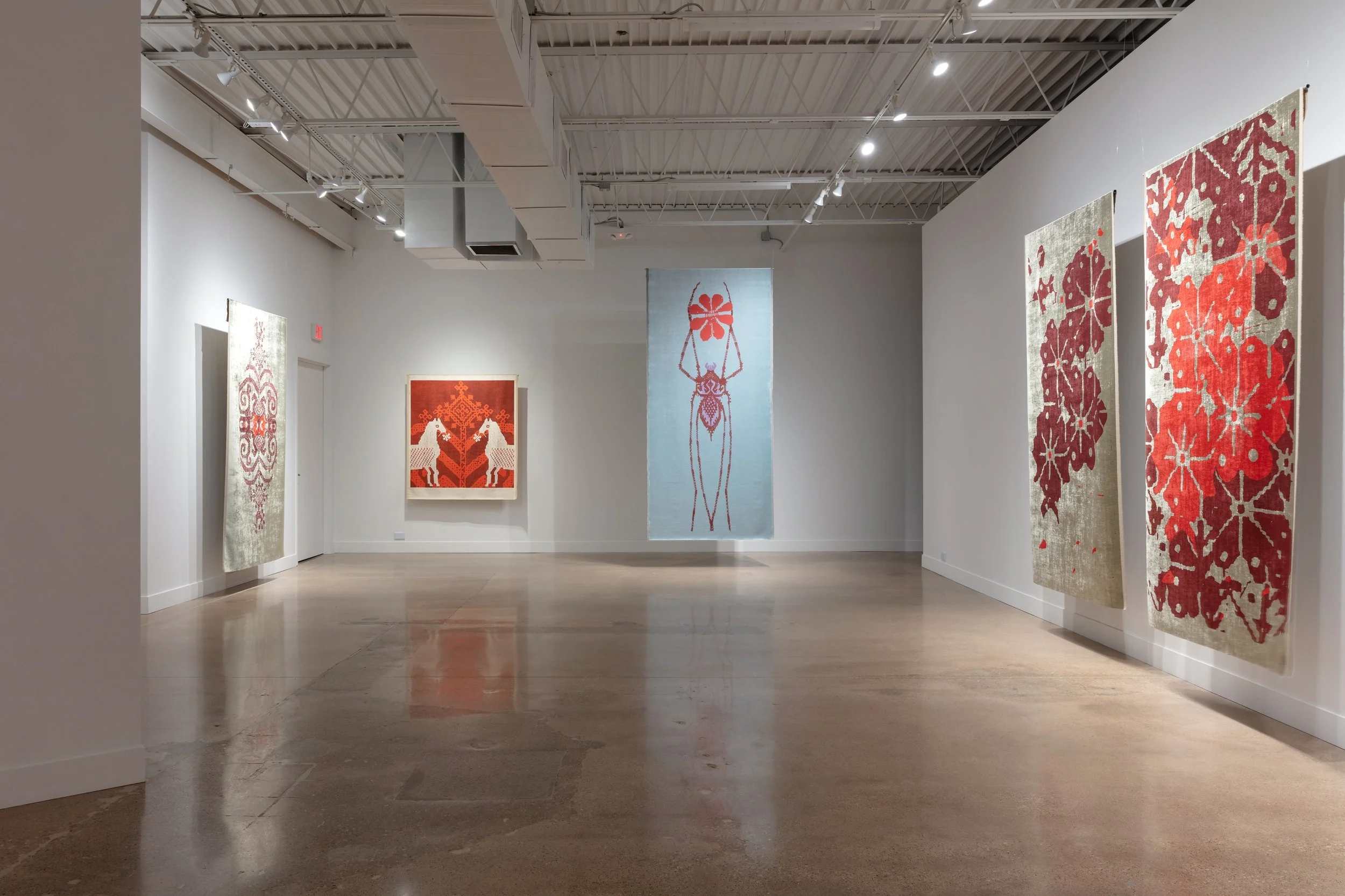 Art gallery with white walls and four large textile art pieces featuring red, beige, and blue colors, displayed on the walls. The gallery has a polished concrete floor and ceiling-mounted lighting.