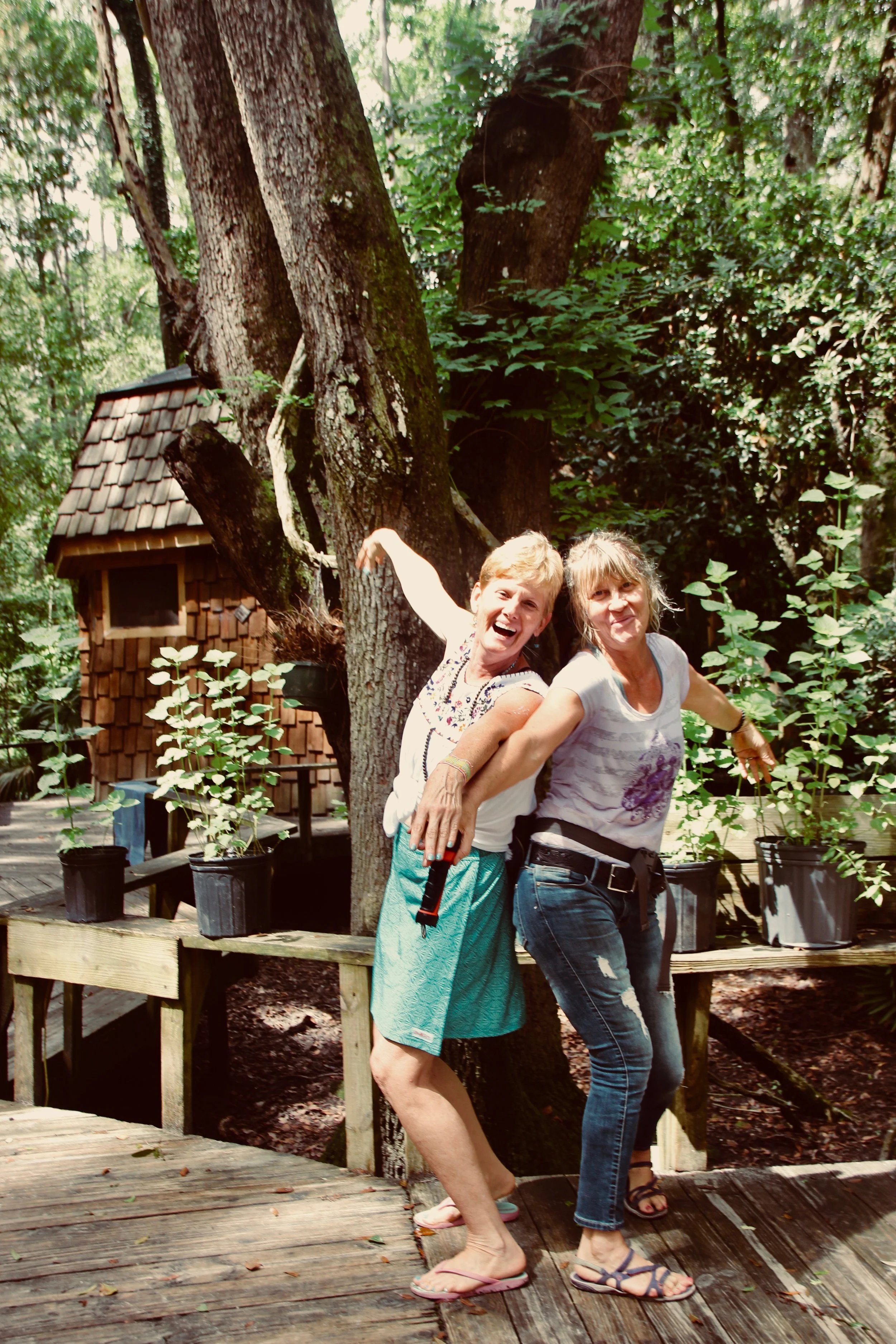 Two people smiling and posing playfully outside in a wooded area, standing on a wooden deck with potted plants around them. A small wooden structure and large trees are visible in the background.