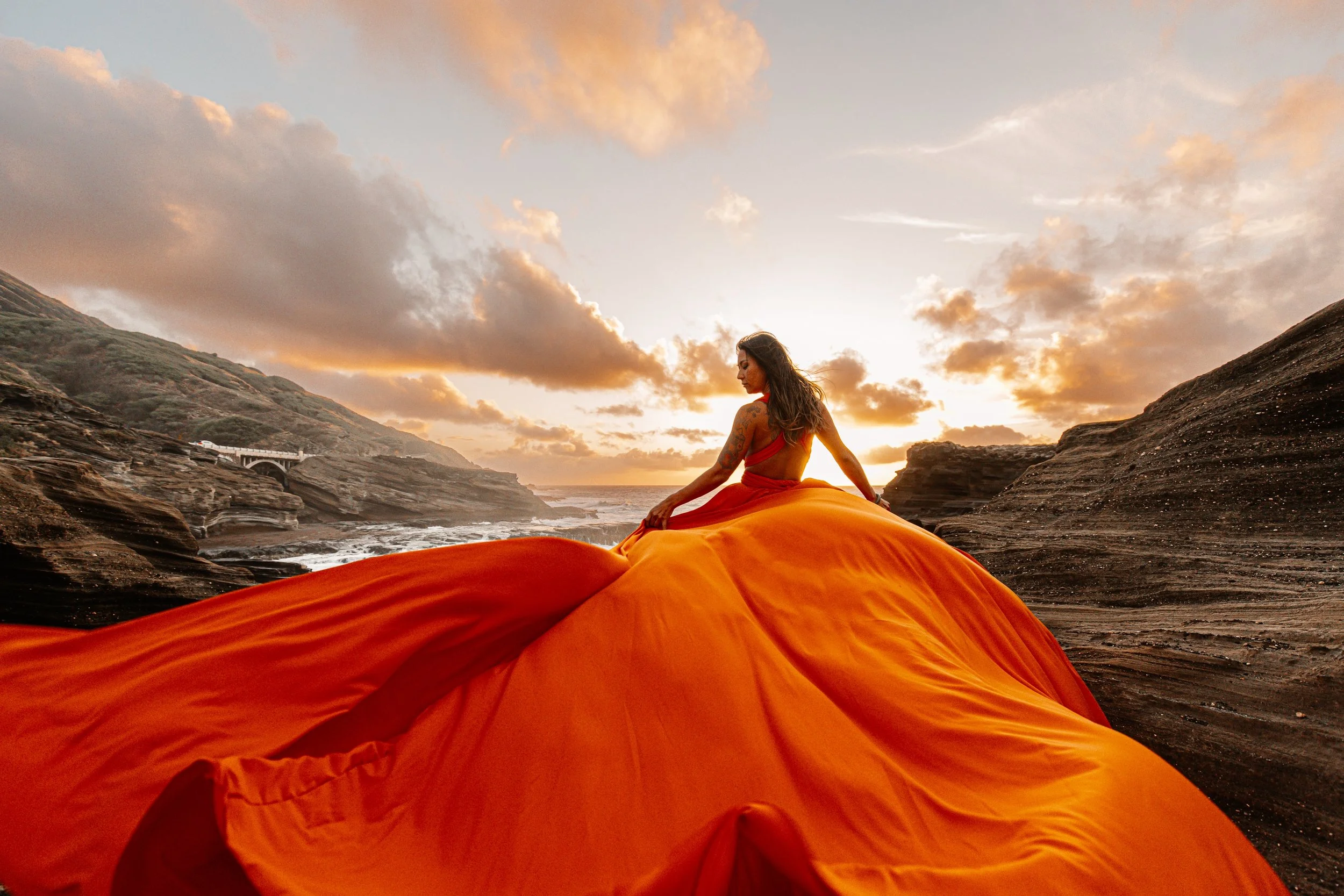 A woman in a flowing orange Hi Flying Dress sitting on rocks at a beach during sunset, with clouds and distant coastline in the background in Maui, Hawaii.