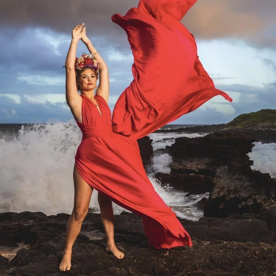 Woman in a flowing red Hi Flying Dress with a floral crown posing on rocky coast with waves crashing behind her in Maui, Hawaii.