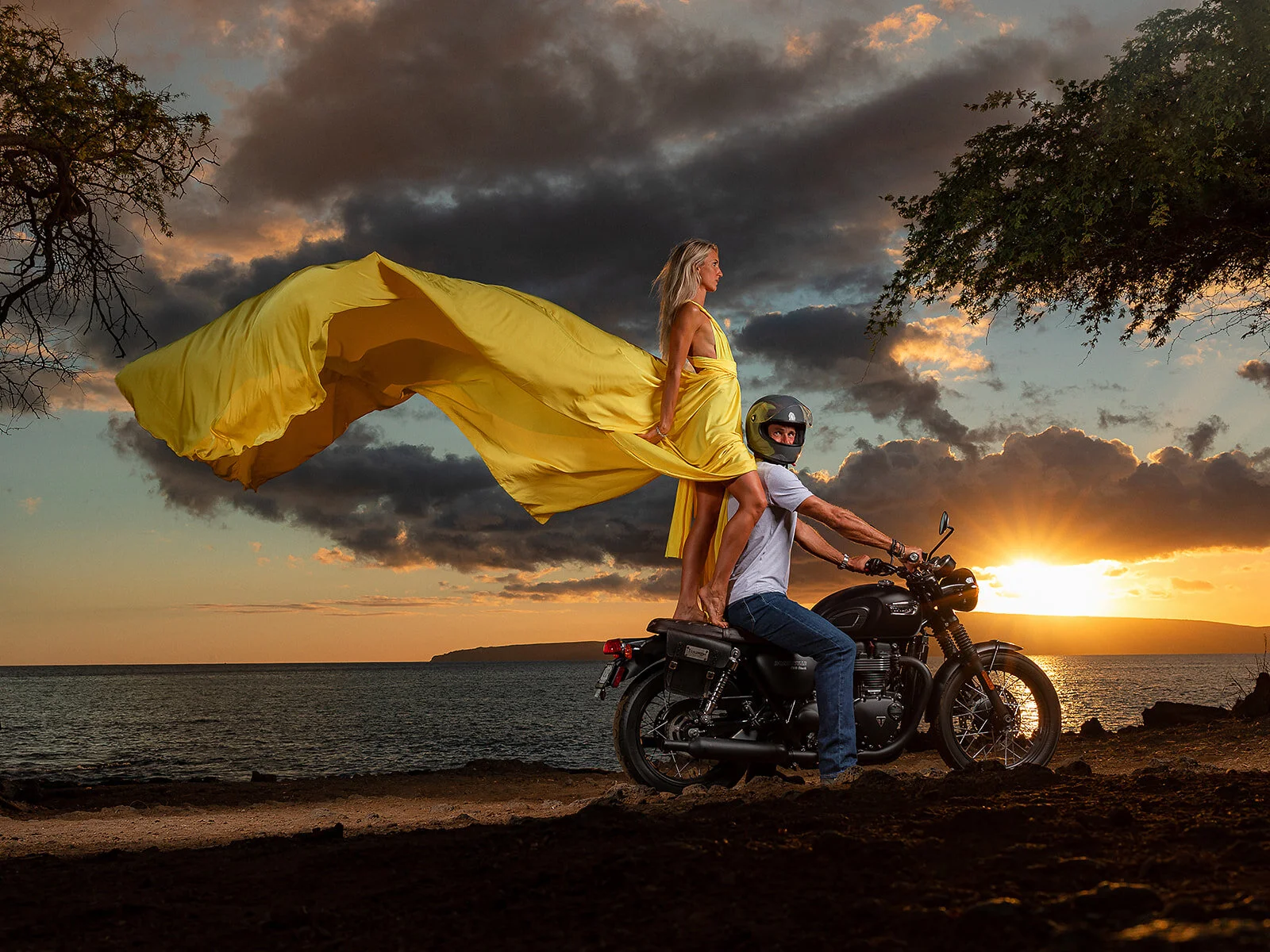 A woman in a flowing yellow Hi Flying Dress riding on the back of a man in a white shirt and jeans, wearing a helmet, on a motorcycle along a beach at sunset with dark clouds in the sky in Maui, Hawaii.