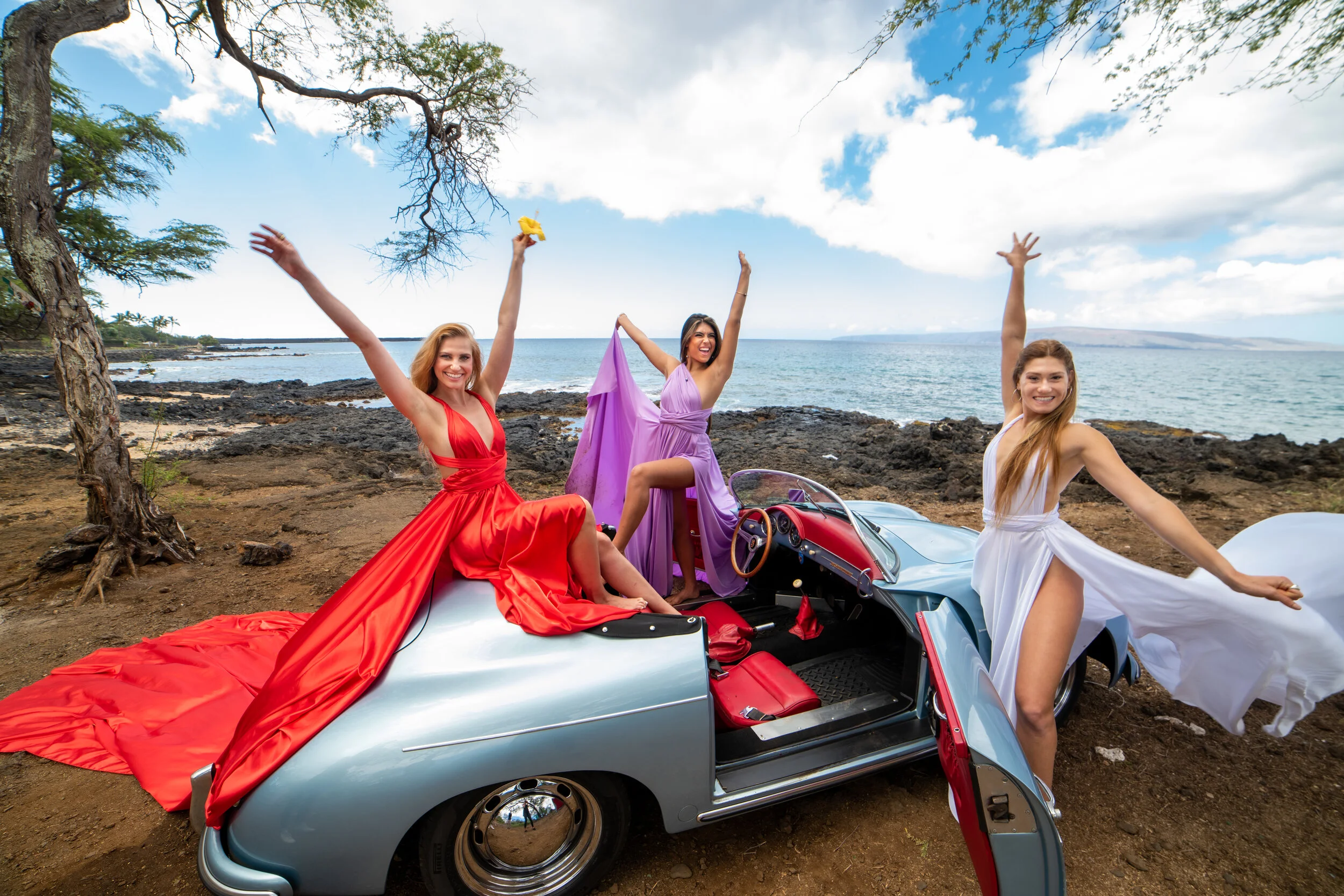Three women in colorful Hi Flying Dresses on a vintage car by the beach, celebrating with applause and joy in Maui, Hawaii.