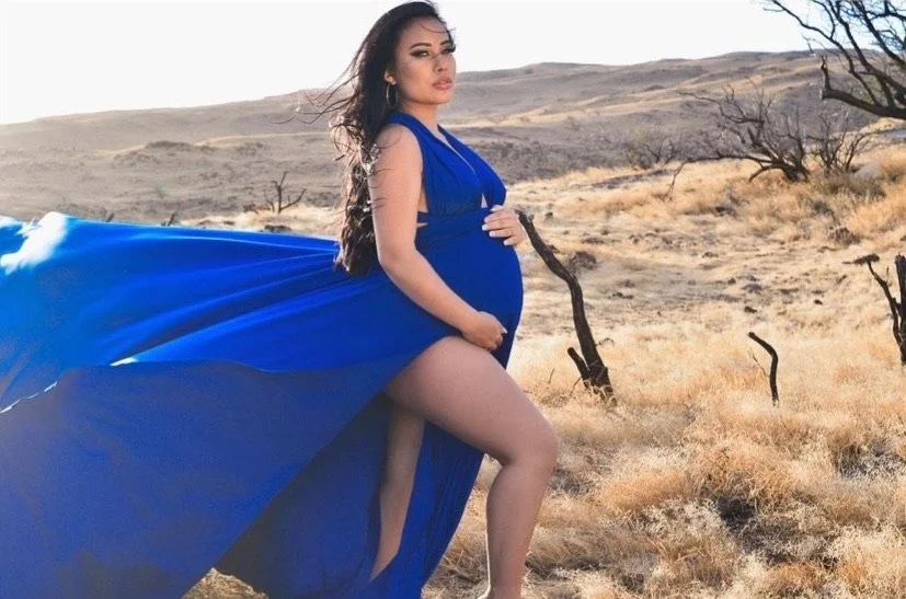 Pregnant woman in a blue Hi Flying Dress standing in a dry, desert landscape with sparse trees in Maui, Hawaii.