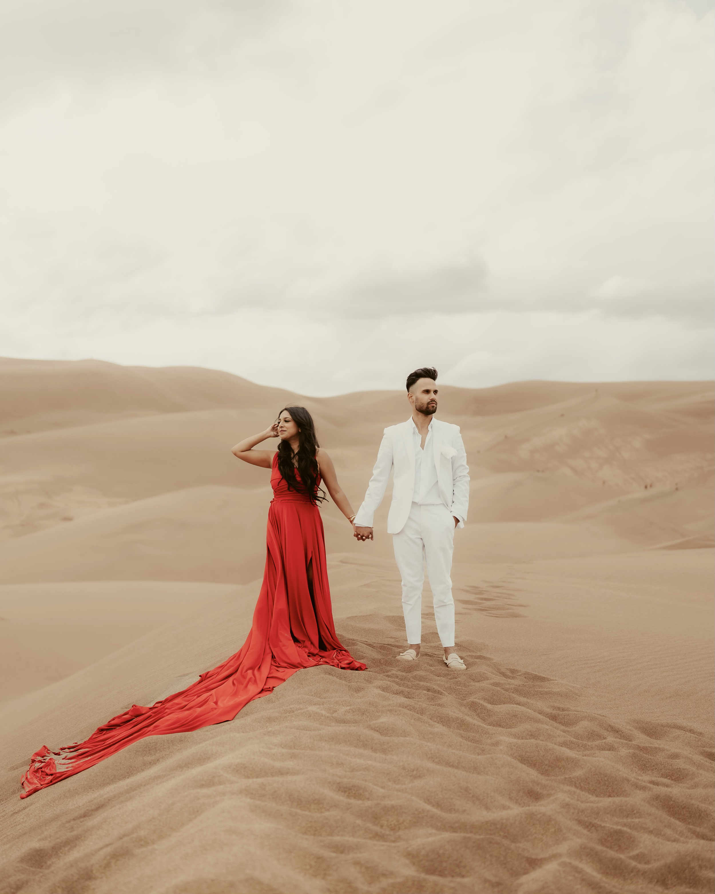 A couple holding hands in a desert landscape, the woman in a long red Hi Flying Dress and the man in a white suit in Dubai.