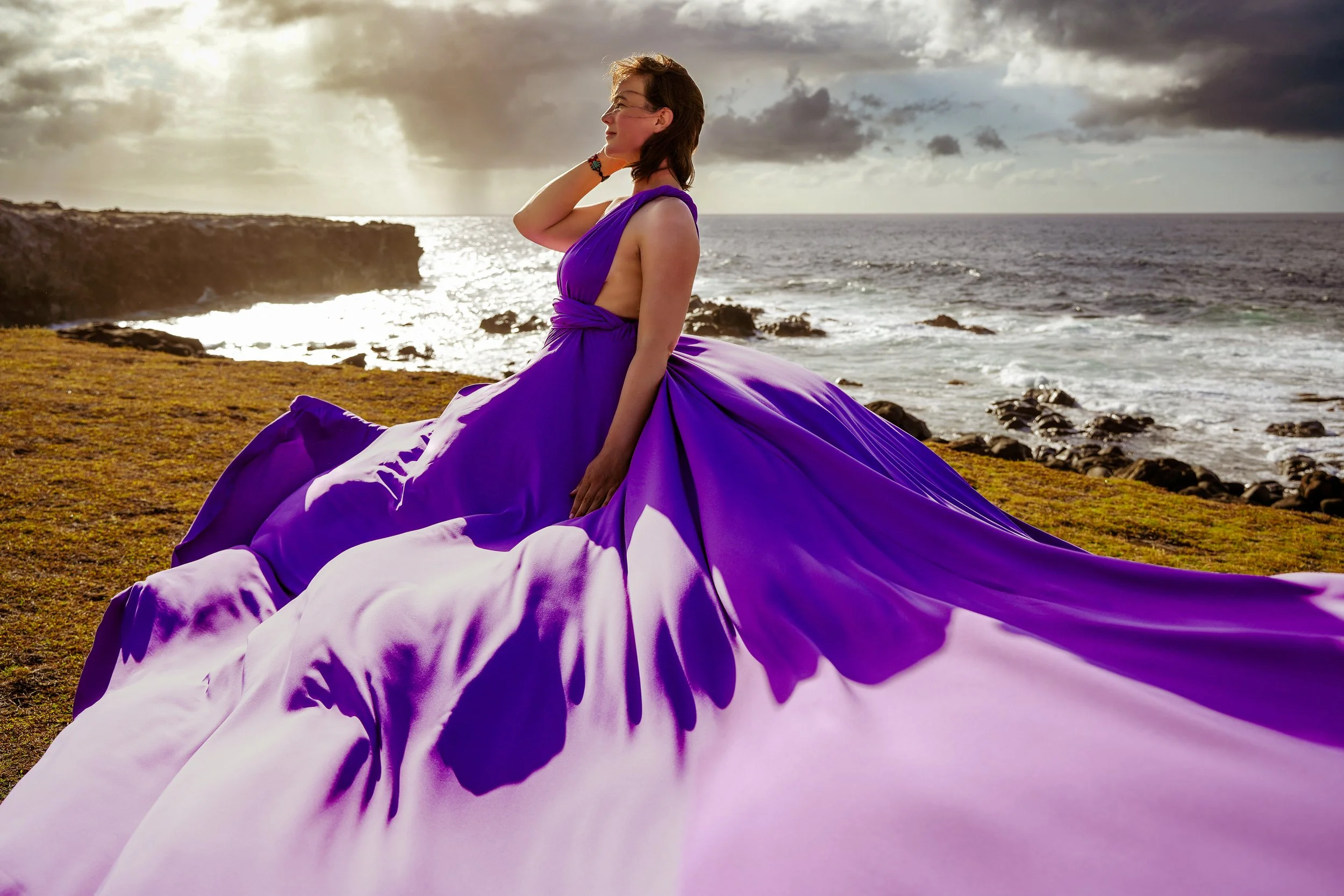 A woman in a flying purple dress sitting on a grassy cliff near the maui ocean at sunset.
