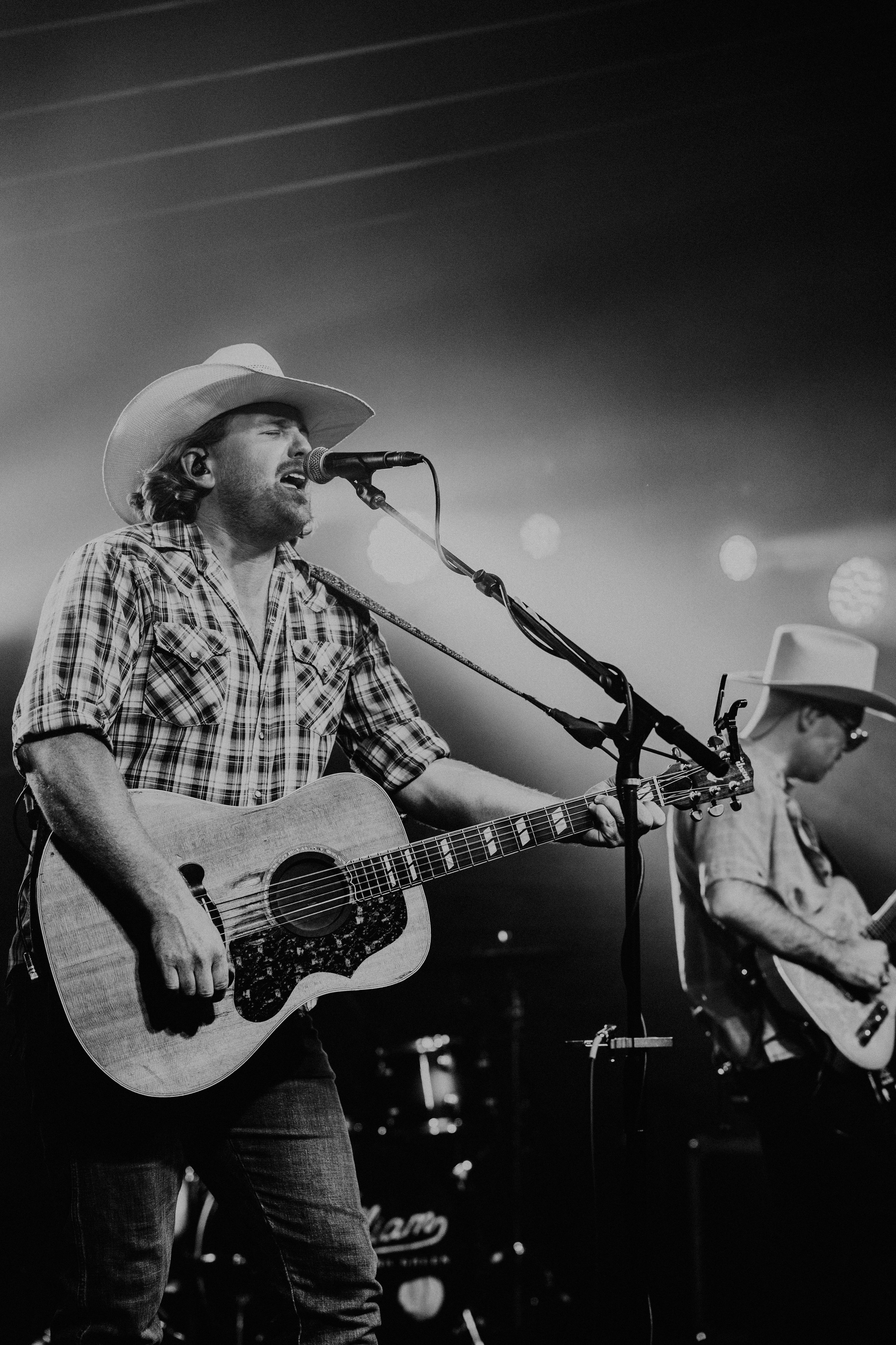 A man in a plaid shirt and cowboy hat singing into a microphone while playing an acoustic guitar on stage, with another musician in the background playing a guitar.