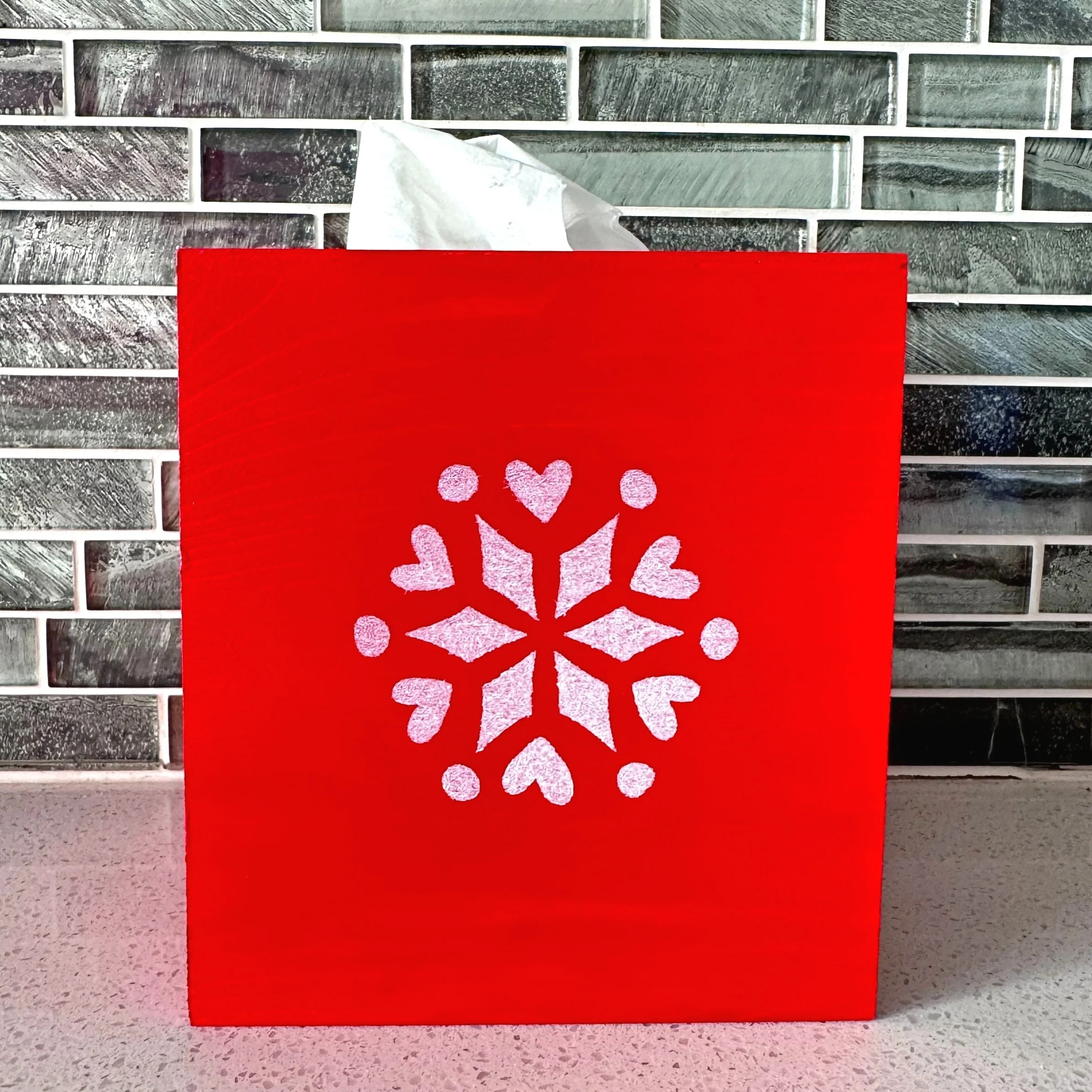 Red gift bag with white snowflake and hearts design, with tissue paper peeking out from the top, on a speckled countertop against a tile backsplash.