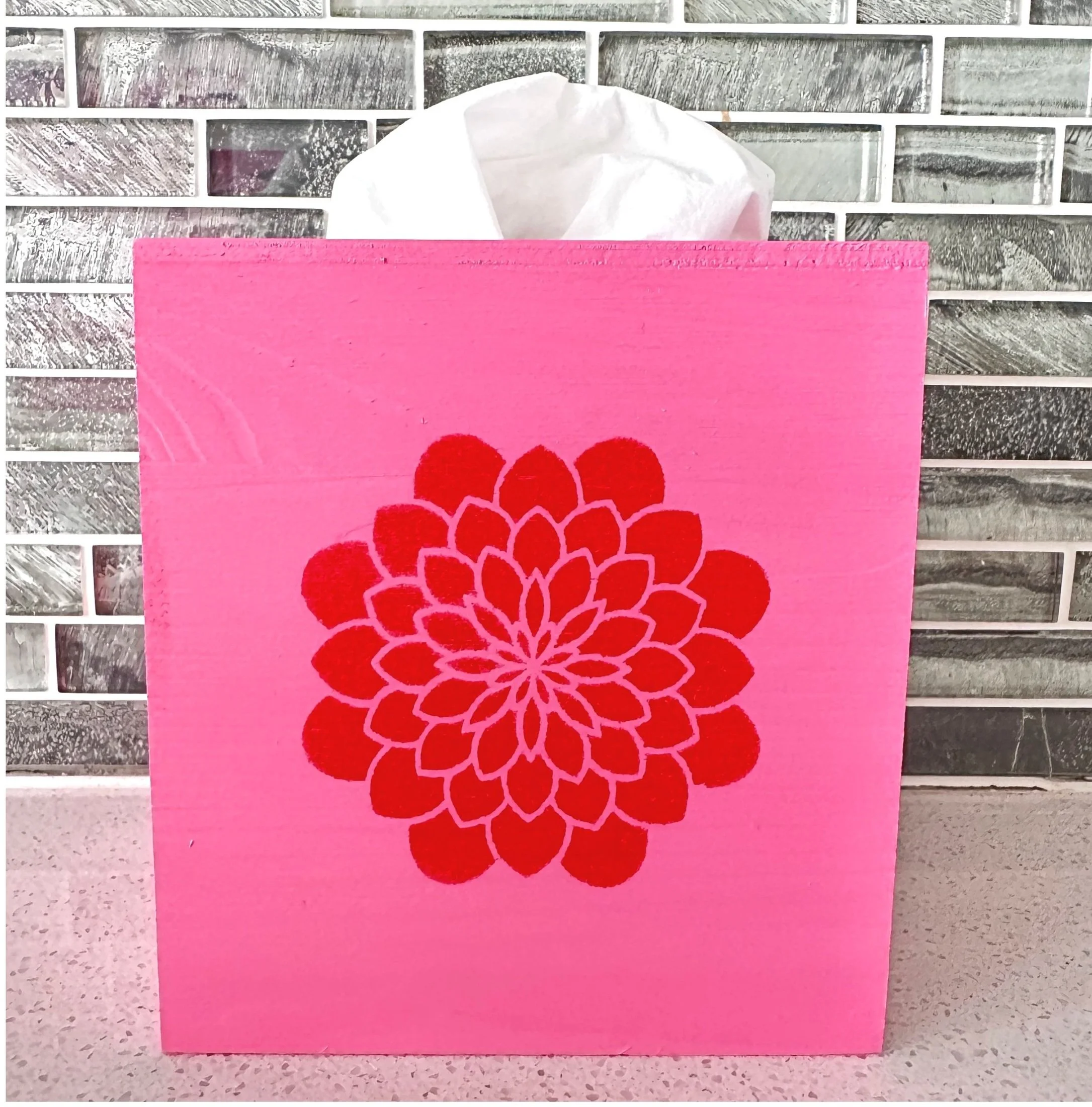 Pink tissue box with a red floral design on its front, with tissue paper sticking out from the top, placed on a light-colored countertop with a gray tiled wall in the background.