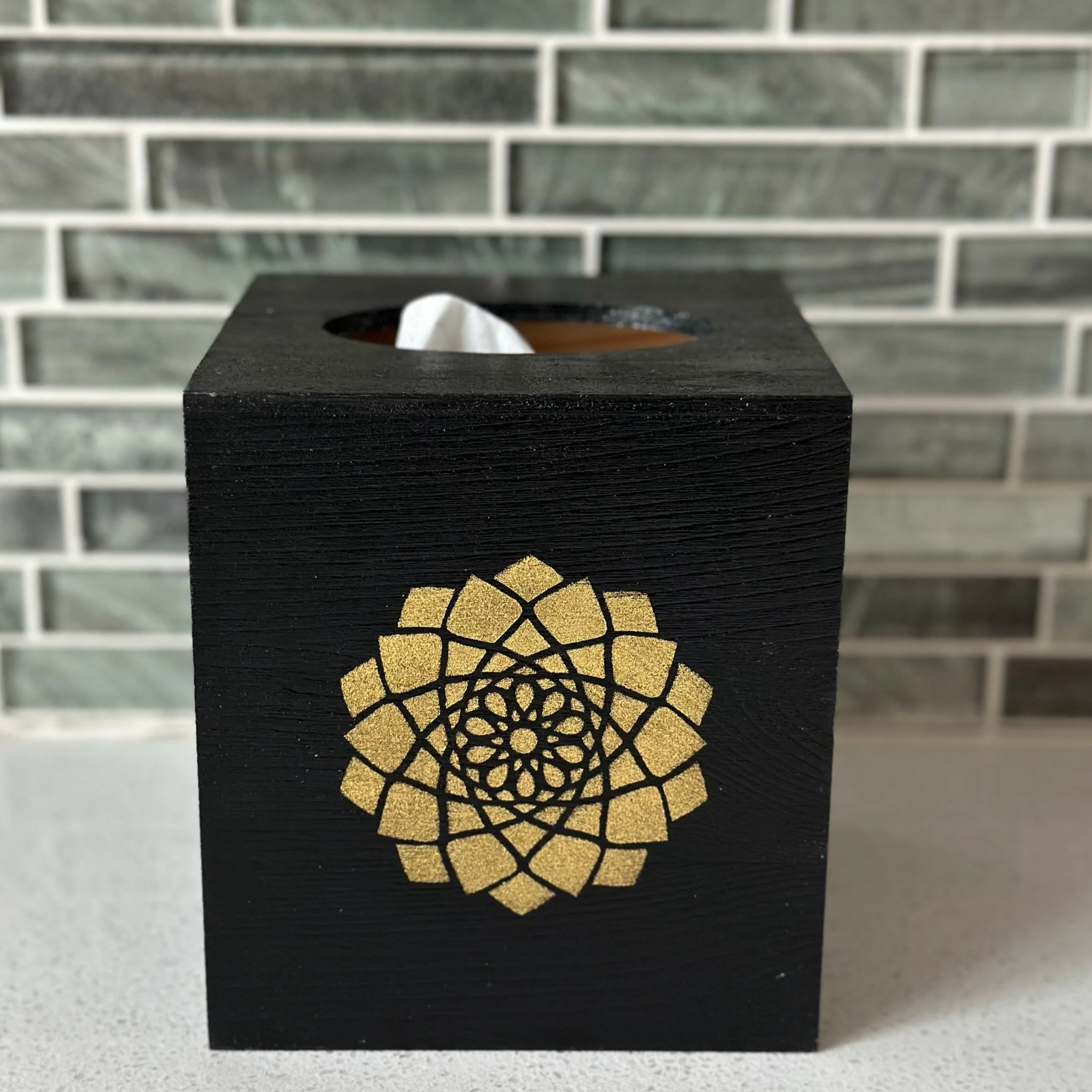 Black tissue box with gold geometric floral design, placed on a light-colored surface, with a gray and white tiled wall in the background.
