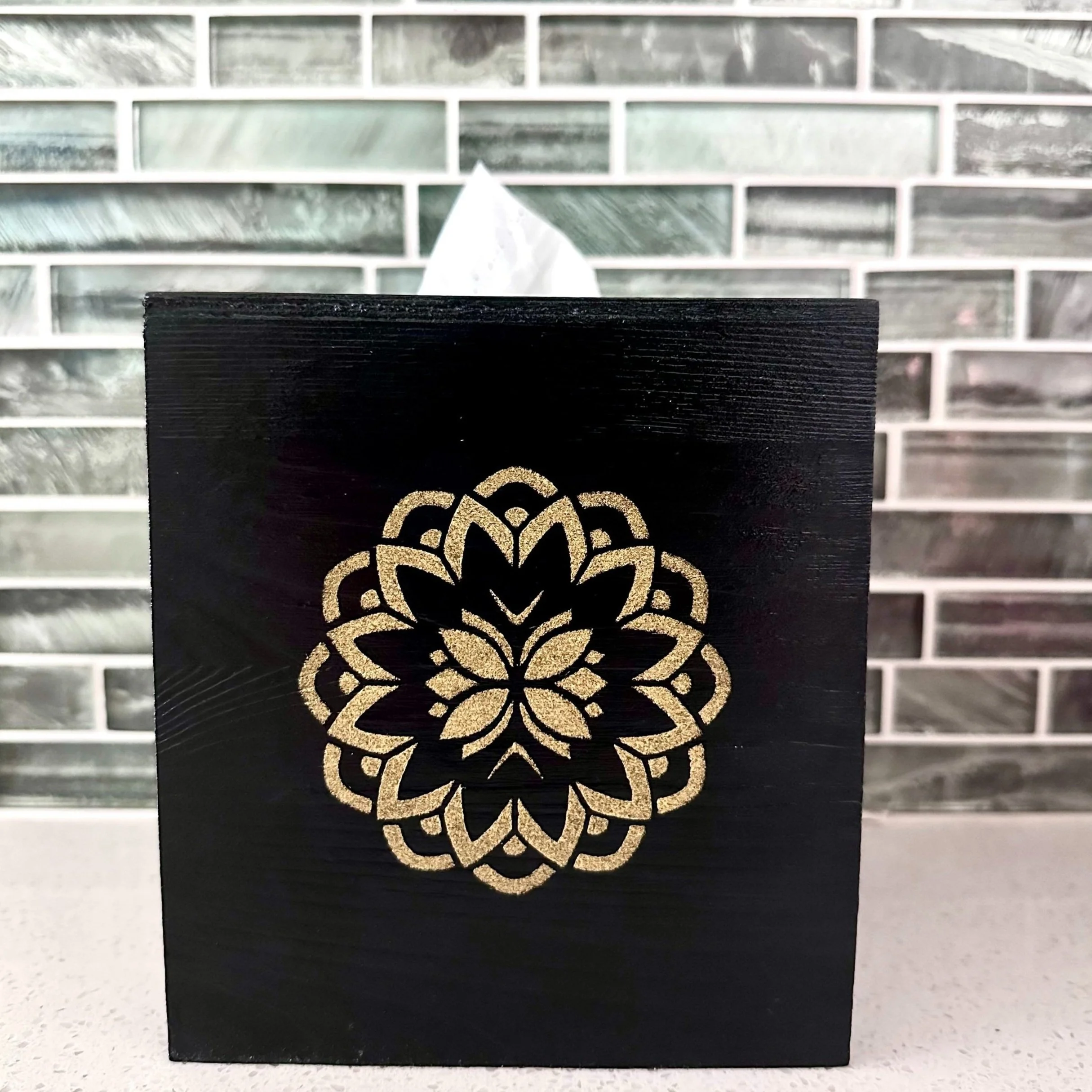 Black box with a gold ornamental floral design on the front, placed on a white countertop with a gray and green tile backsplash in the background.