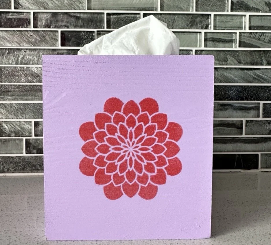 A pink tissue box with a red floral design on the front, with tissues protruding from the top, placed in front of a black and gray tiled backsplash.