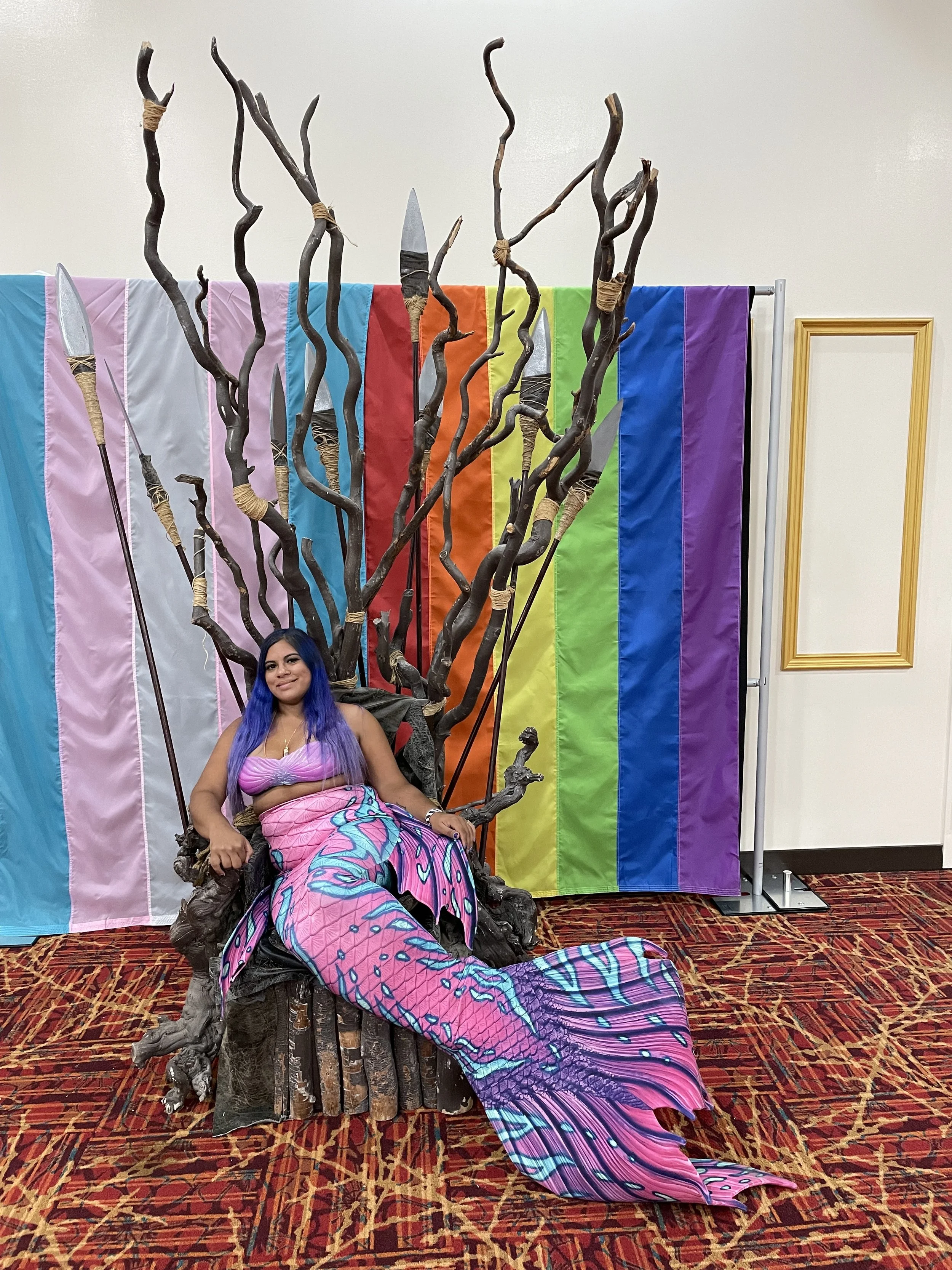 A brown-skinned woman with blue hair sits smiling on Lexa's throne from "The 100" in front of an LGBTQ+ flag wearing a pink mermaid costume with electric blue designs on the tail.