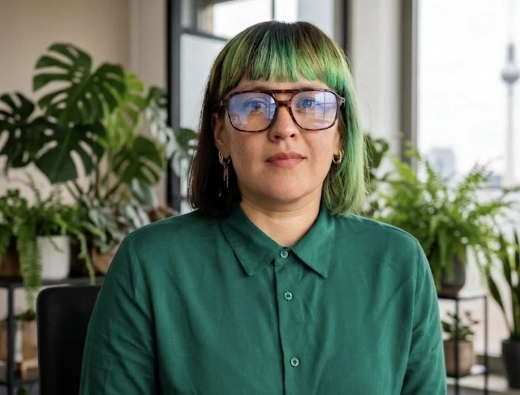 A woman with green and black hair, wearing glasses and a green collared shirt, sitting in front of a window with potted plants around her.