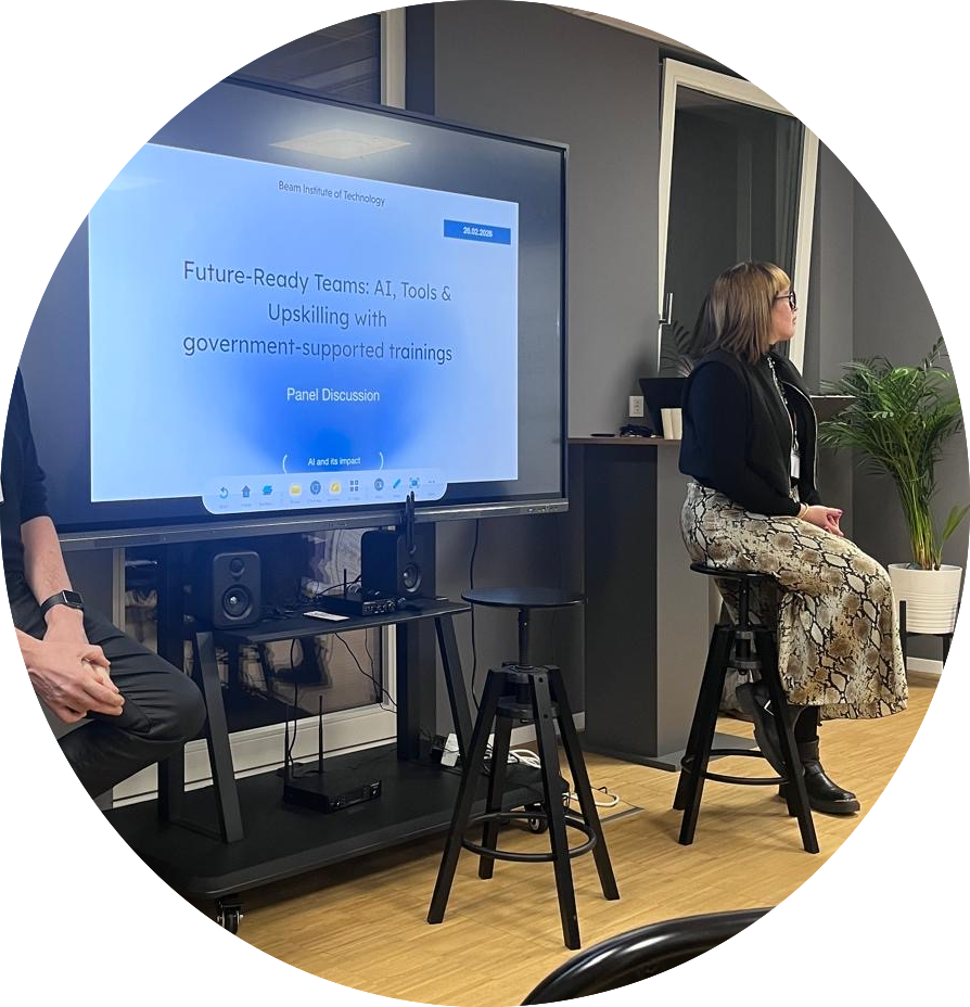 Woman sitting on a stool in front of a large monitor during a panel discussion about AI, tools, and upskilling at a government-supported training event.