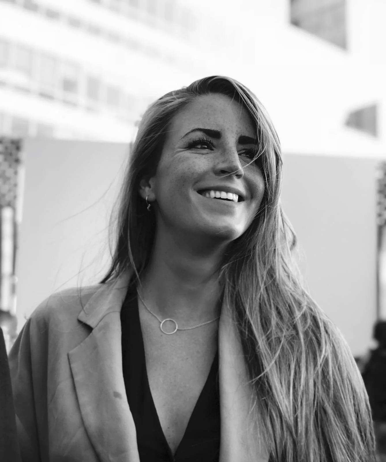 Black and white photo of a smiling woman with long hair, wearing a necklace, earrings, and a light-colored jacket.