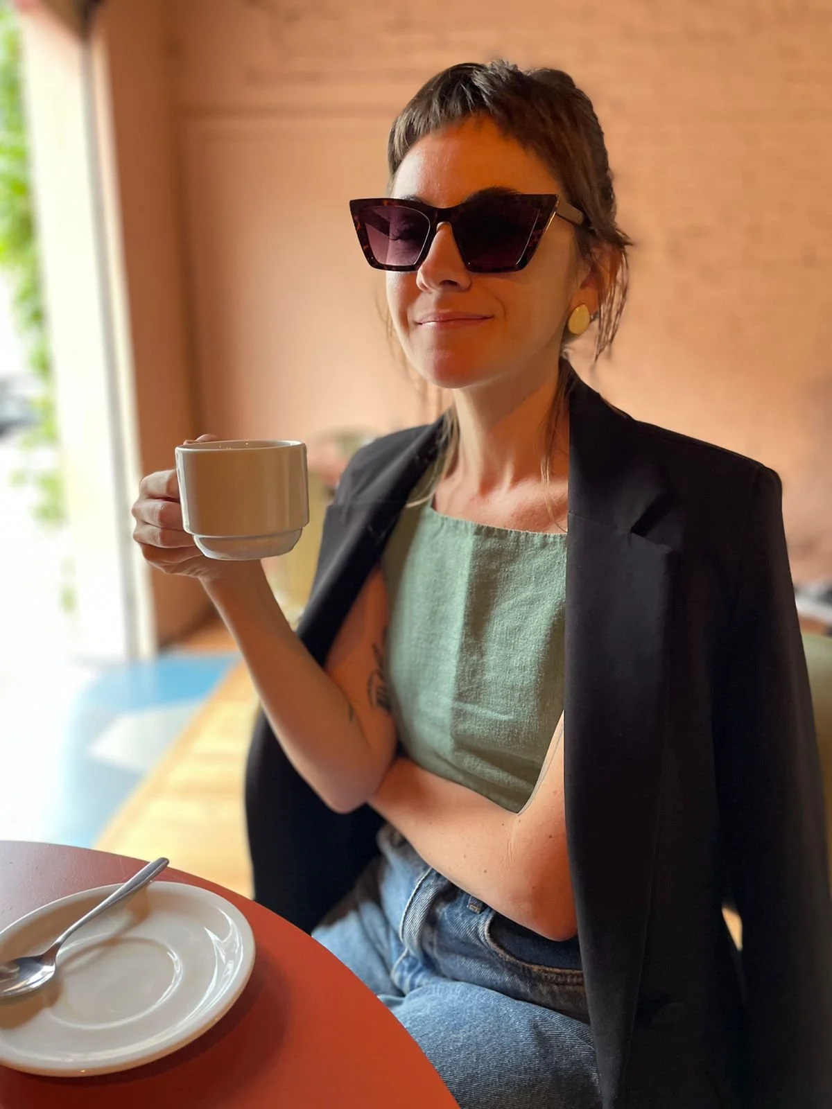 Woman with short hair wearing sunglasses, a green top, and a black blazer, sitting at a table with a white cup in her hand, a plate with a spoon on it in front of her, and smiling.