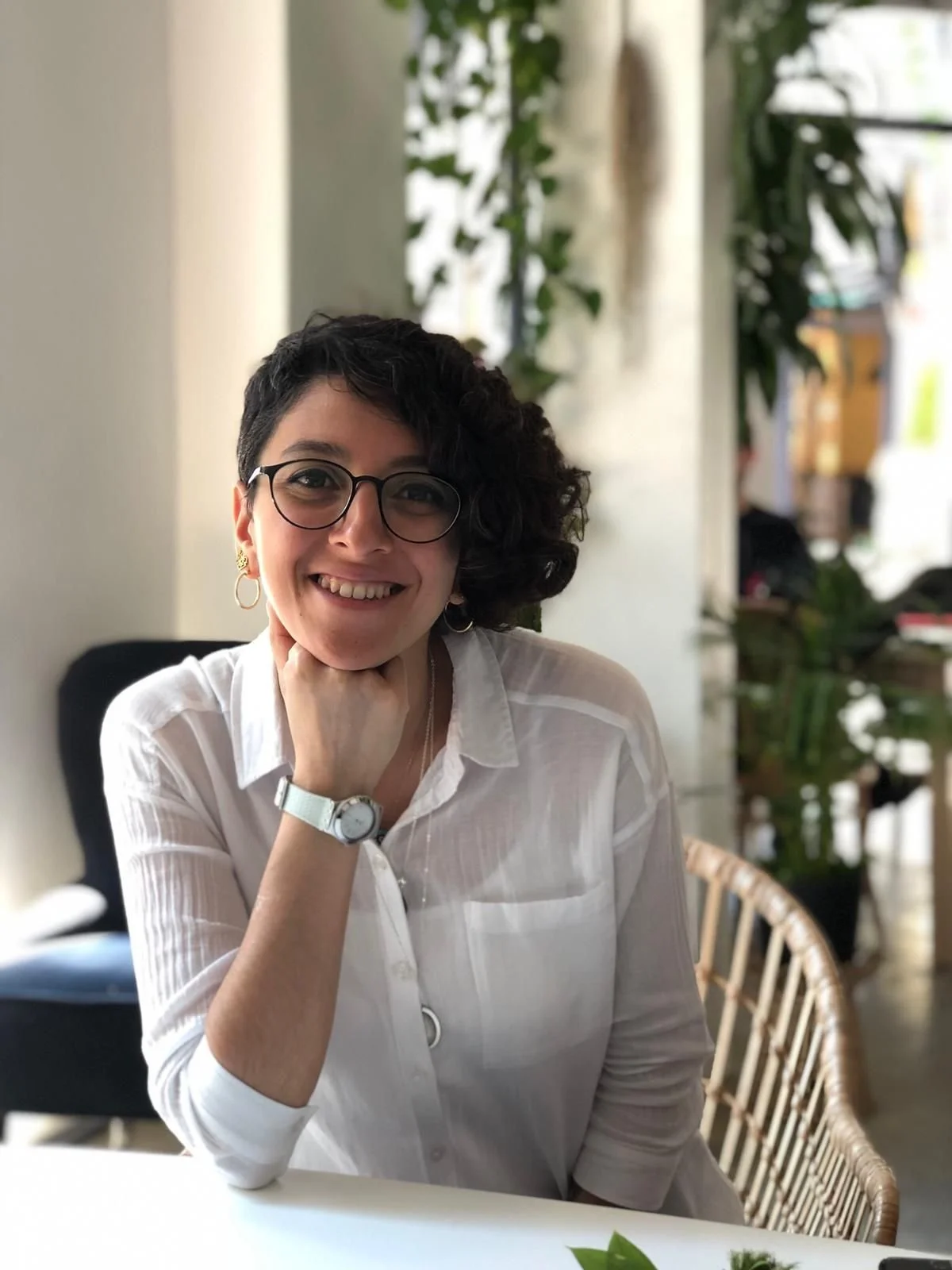 A woman with short curly dark hair and glasses, wearing a white shirt, smiling and resting her chin on her hand, sitting at a table in a well-lit indoor space with plants and a wicker chair in the background.
