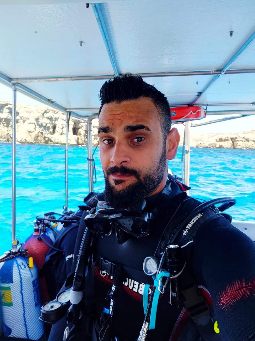 A man in scuba diving gear on a boat with blue water and rocky cliffs in the background.