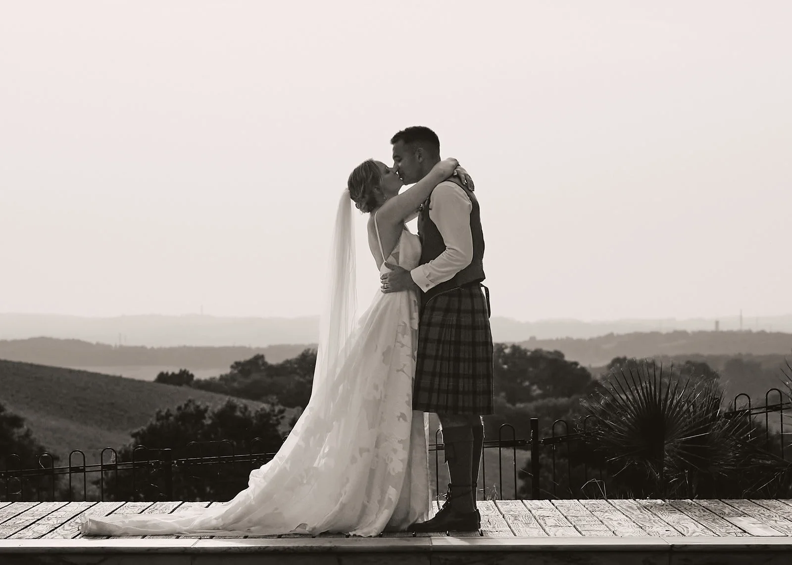 A bride and groom passionately kiss on a wooden platform with scenic rolling hills in the background, black and white photo.
