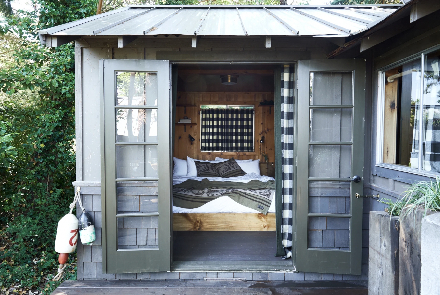 A tiny house with open double doors, revealing a bed inside and surrounded by greenery, with buoys hanging outside.