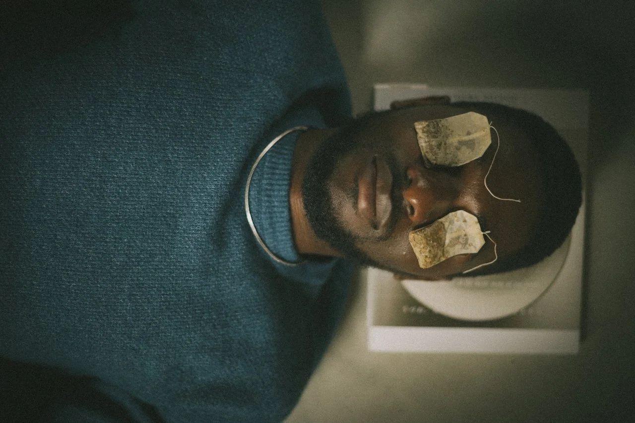 A man with closed eyes and a beard lying down with two tea bags on his eyelids, possibly in a spa or relaxation setting.