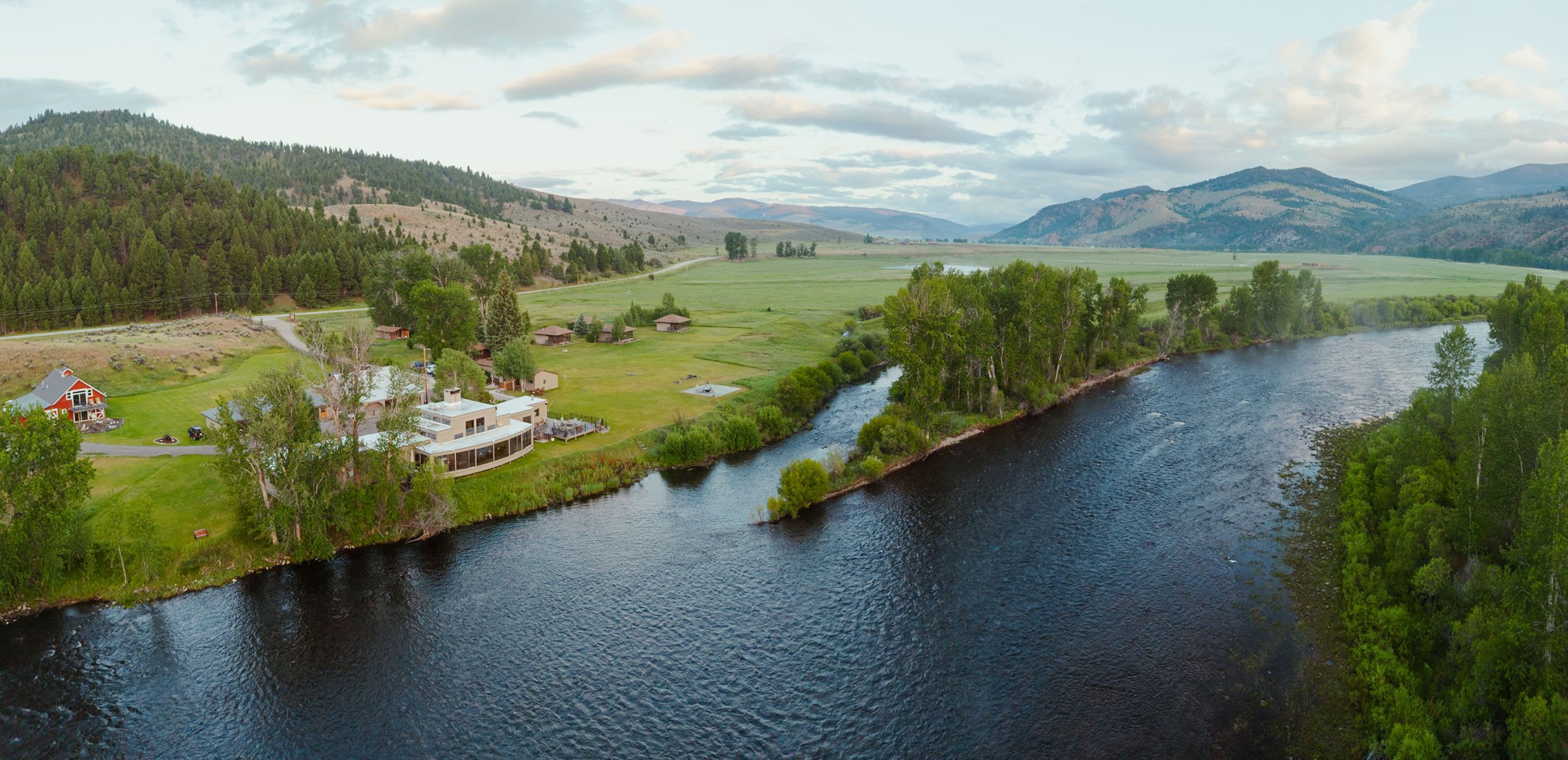 A lodge with big glass windows sits at the edge of the Big Hole River in Montana. The river is in the foreground. Green meadows, mountains, and clouds fill the background.