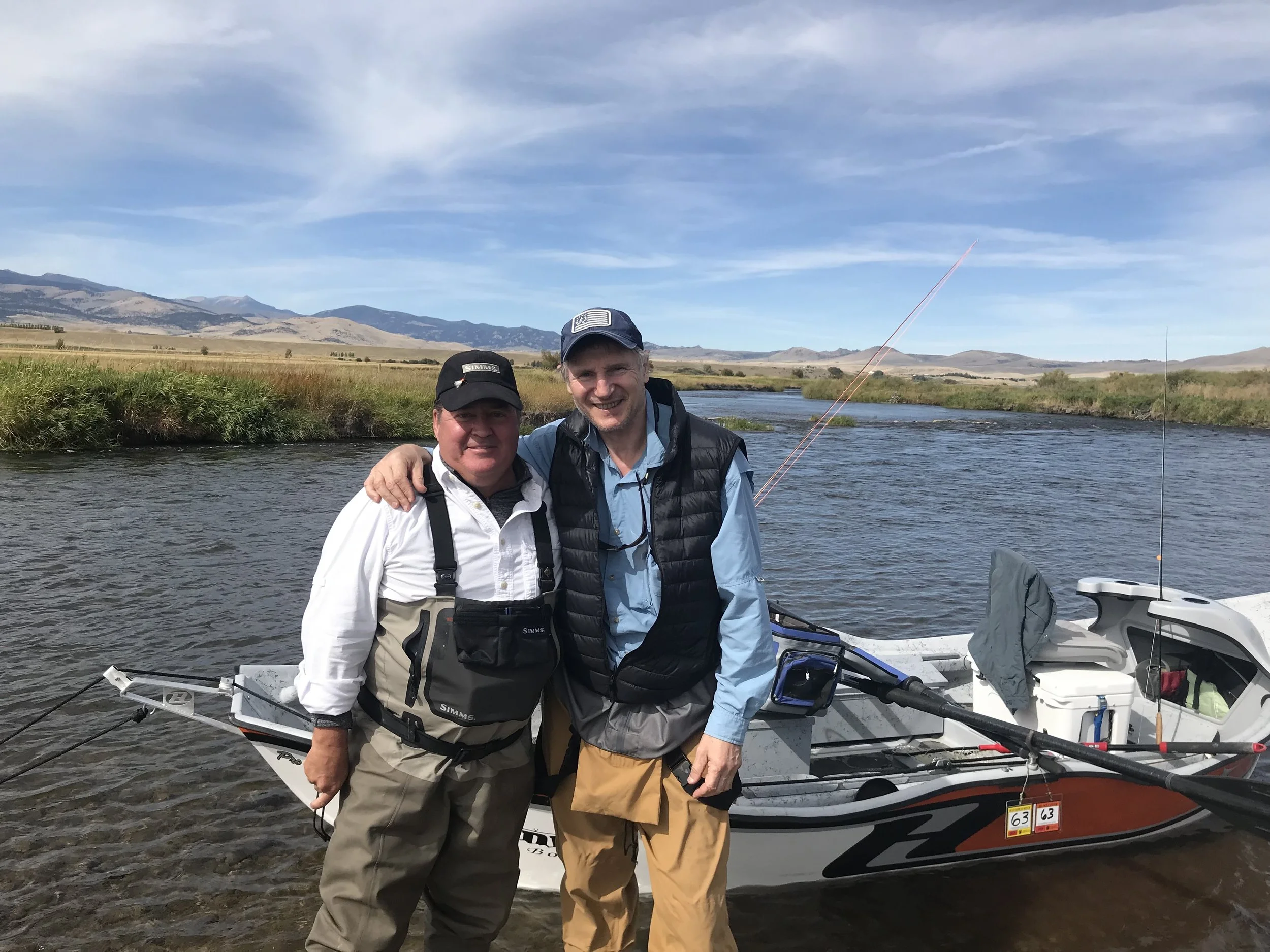 Author John Barrett stands in a river beside actor Liam Neeson. Behind them is a drift boat and a pair of fly rods, and the background shows Montana's classic Big Sky.