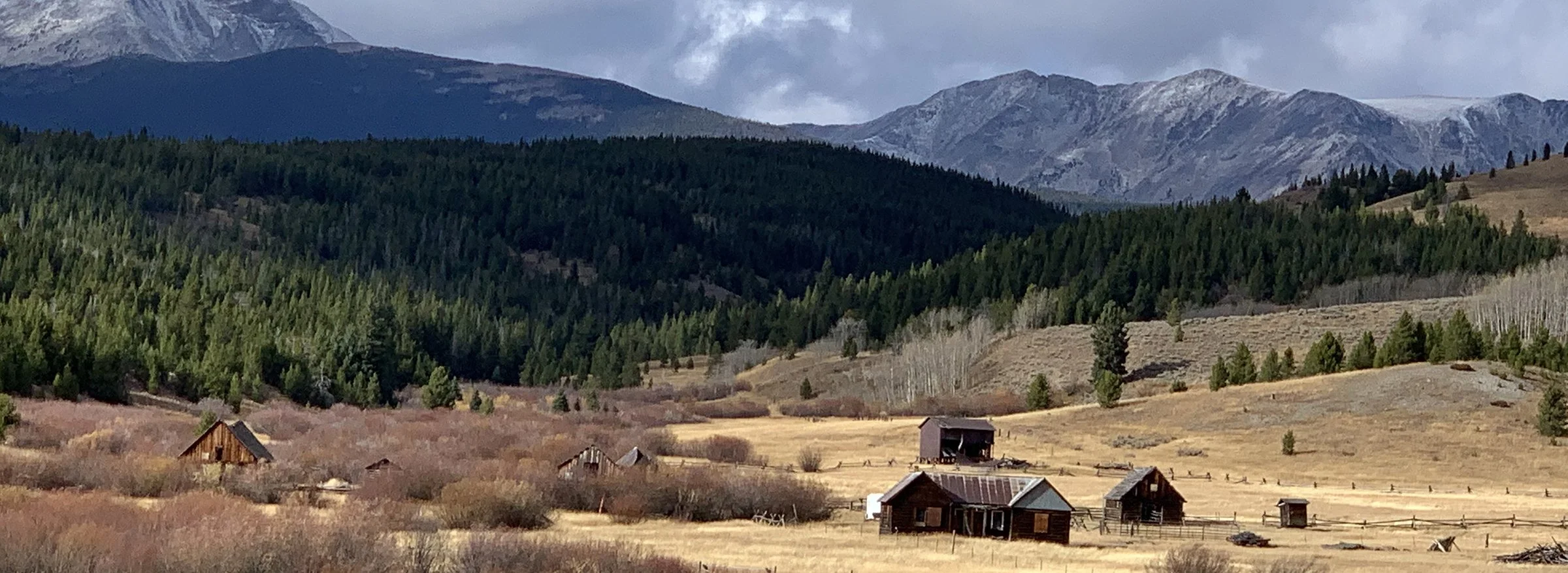 A panoramic view of the upper Big Hole Valley shows aseveral old homestead buildings in the foreground. A thickly forested foothill in the background is framed by massive, snow-dusted granite peaks in the background.