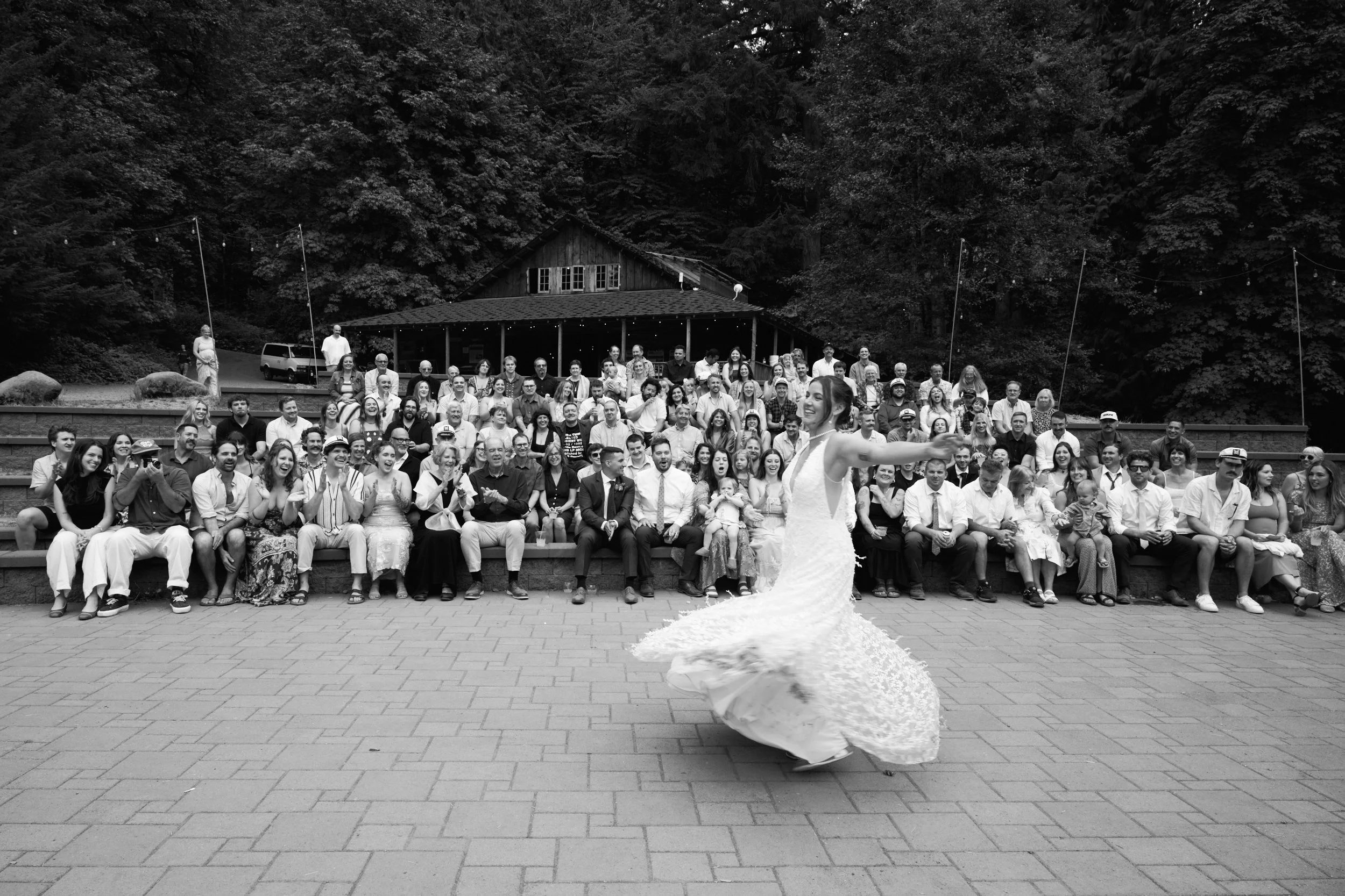 Black and white image of a bride dancing and spinning in front of hundreds of wedding guests and her husband of course as they cheer her on in a forest outside Portland Oregon.