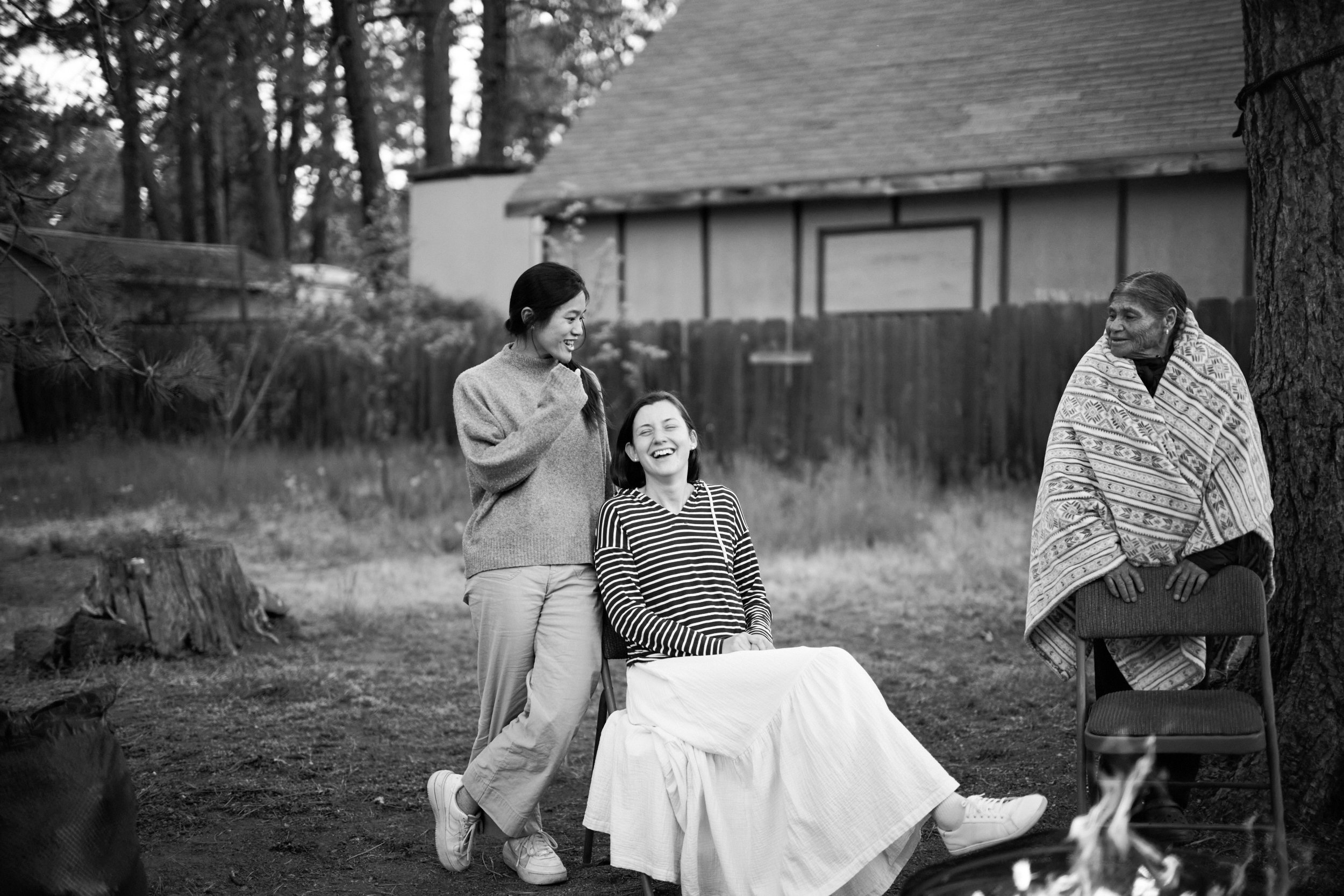 During a rehearsal dinner at their home a bride sits in front of a camp fire laughing with her best friend as the groom grandmother, visiting from Mexico, looks on with a smile.