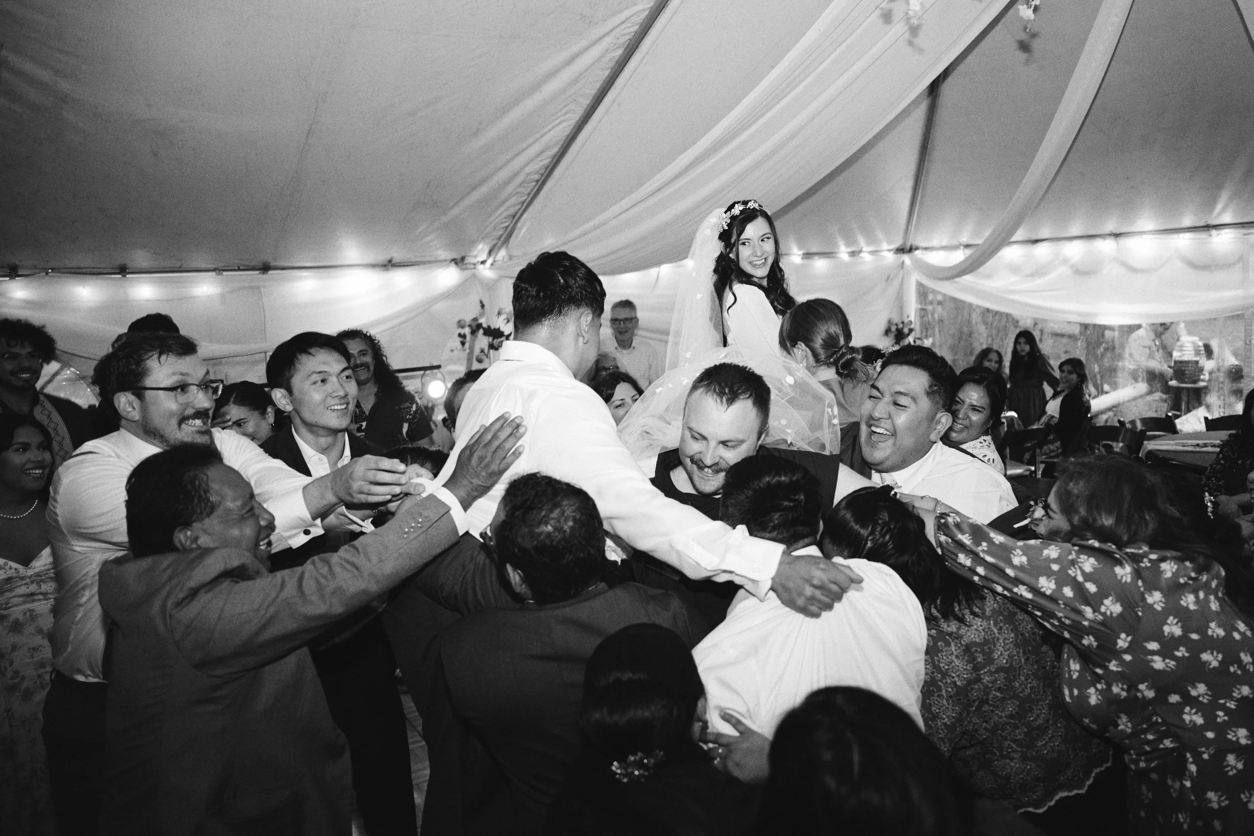 A bride is lifted onto a chair and protected by the women around her while the grooms friends and family try to knock her off the chair. The scene, tucked into the Deschutes National Forest near Bend, Oregon is full of chaos and joy.