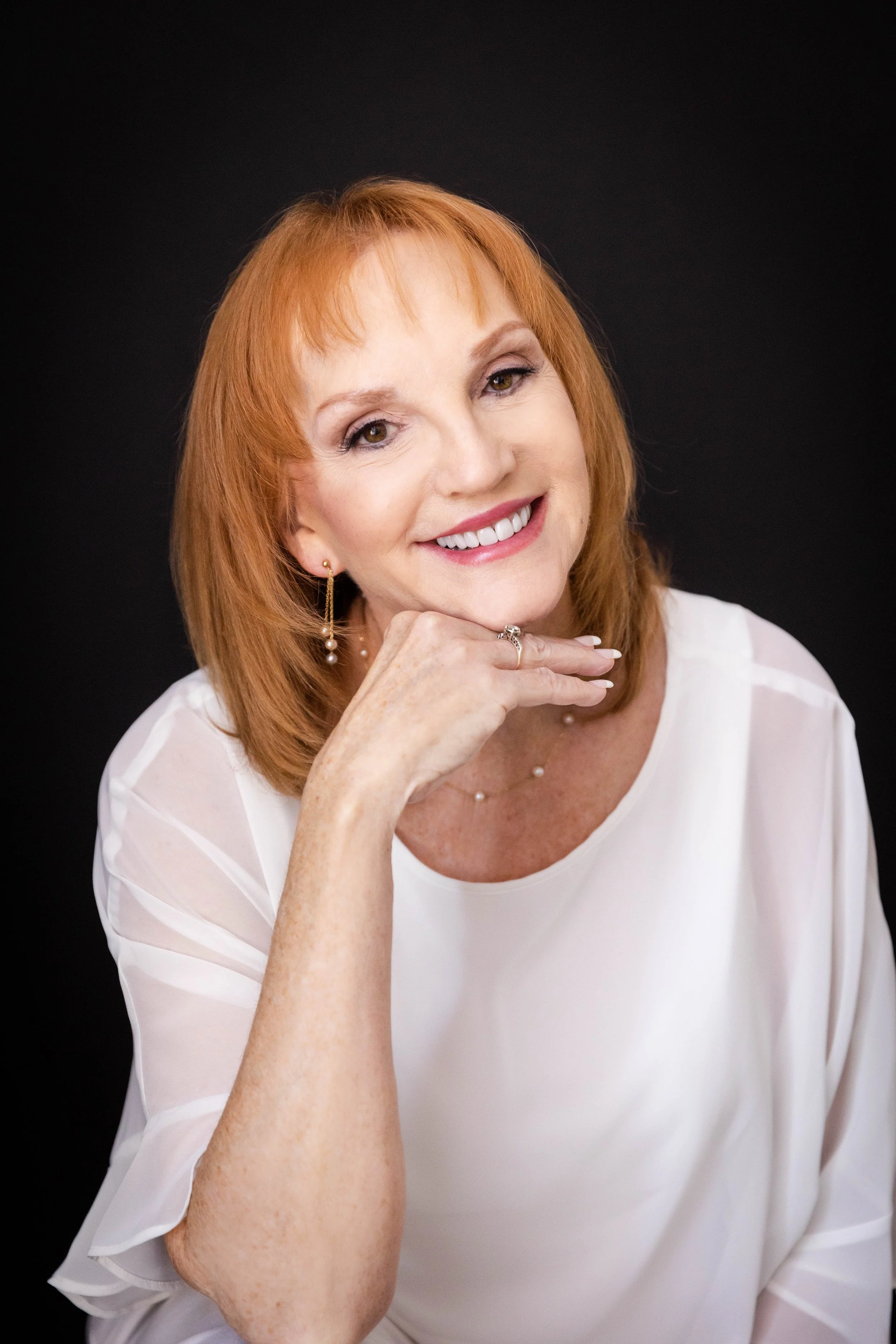 A middle-aged woman with red hair, wearing a white sheer blouse, pearl earrings, a ring, and a pearl necklace, smiling, resting her chin on her hand, against a black background.