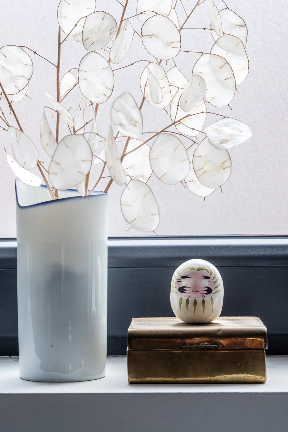 Een witte Piet Stockmans vaas met droogbloemen en een traditionele Japanse darumapop op de vensterbank van de bureau van de Leuvense grafisch ontwerper en Japan-liefhebber Maarten Deckers.