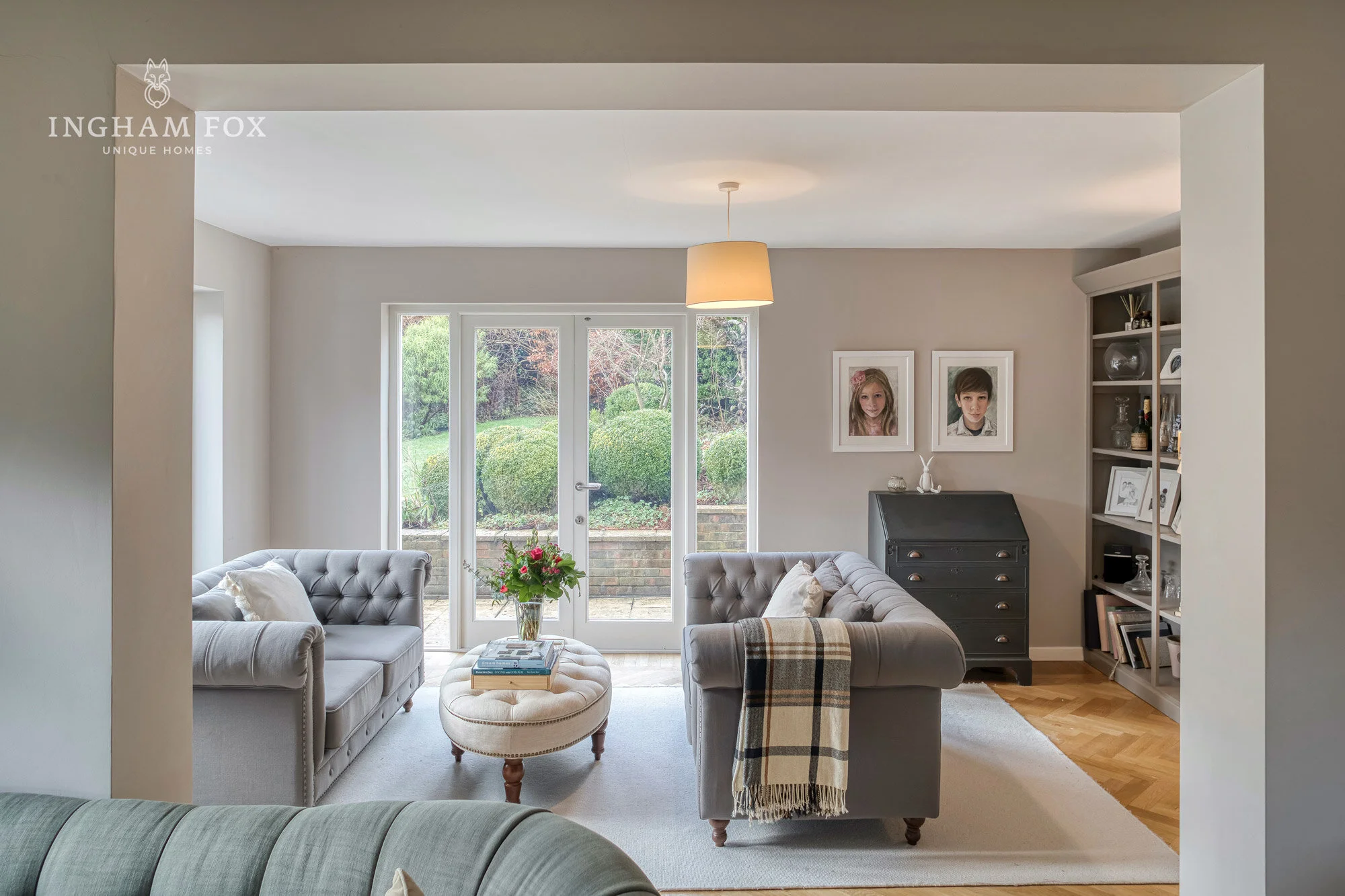 Modern living room with gray sofas, a round ottoman, large windows overlooking a garden, and framed photos on the wall.