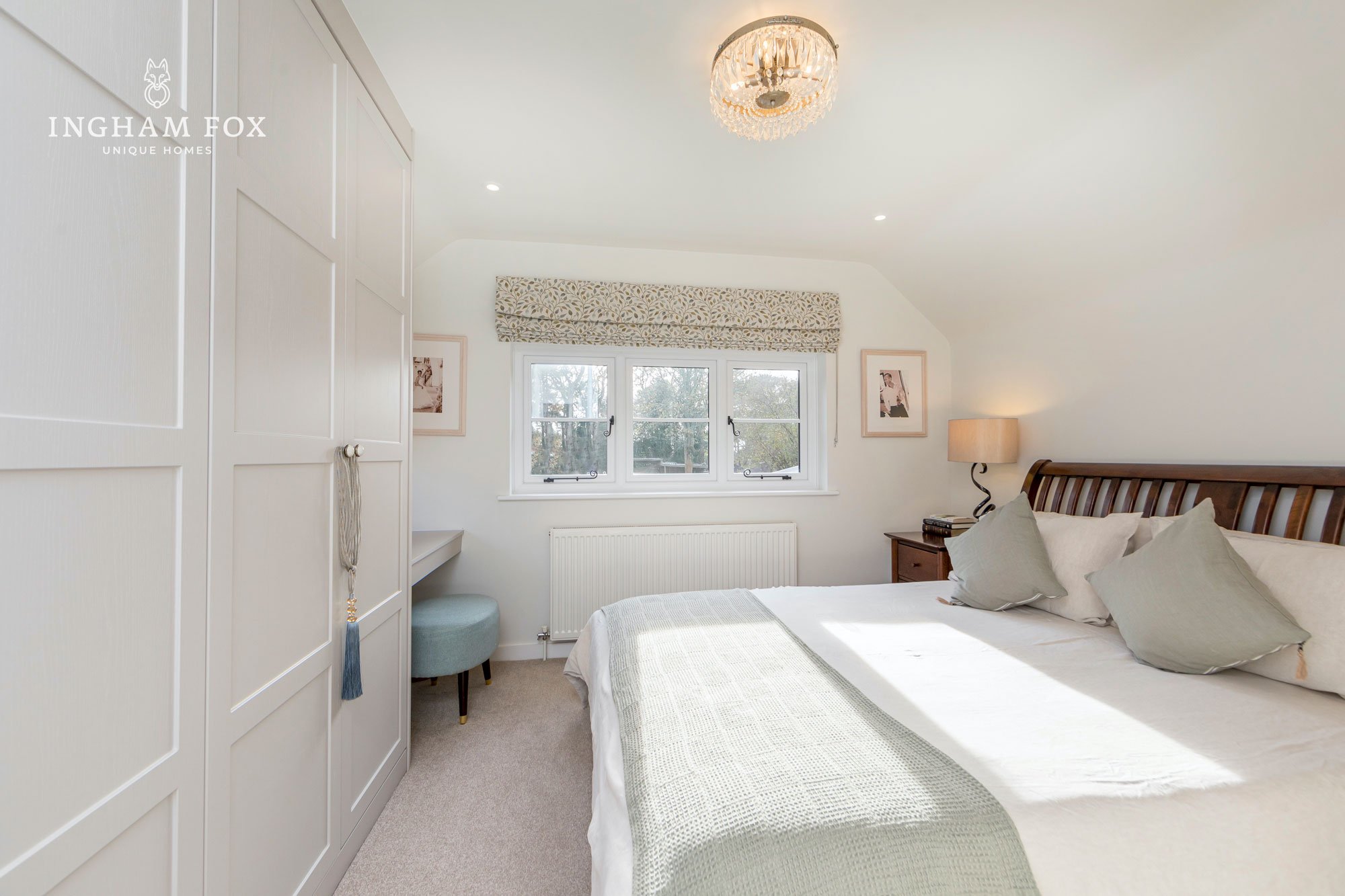 Bright bedroom with a large bed, white walls, decorative chandelier, and sunlight streaming through a window.