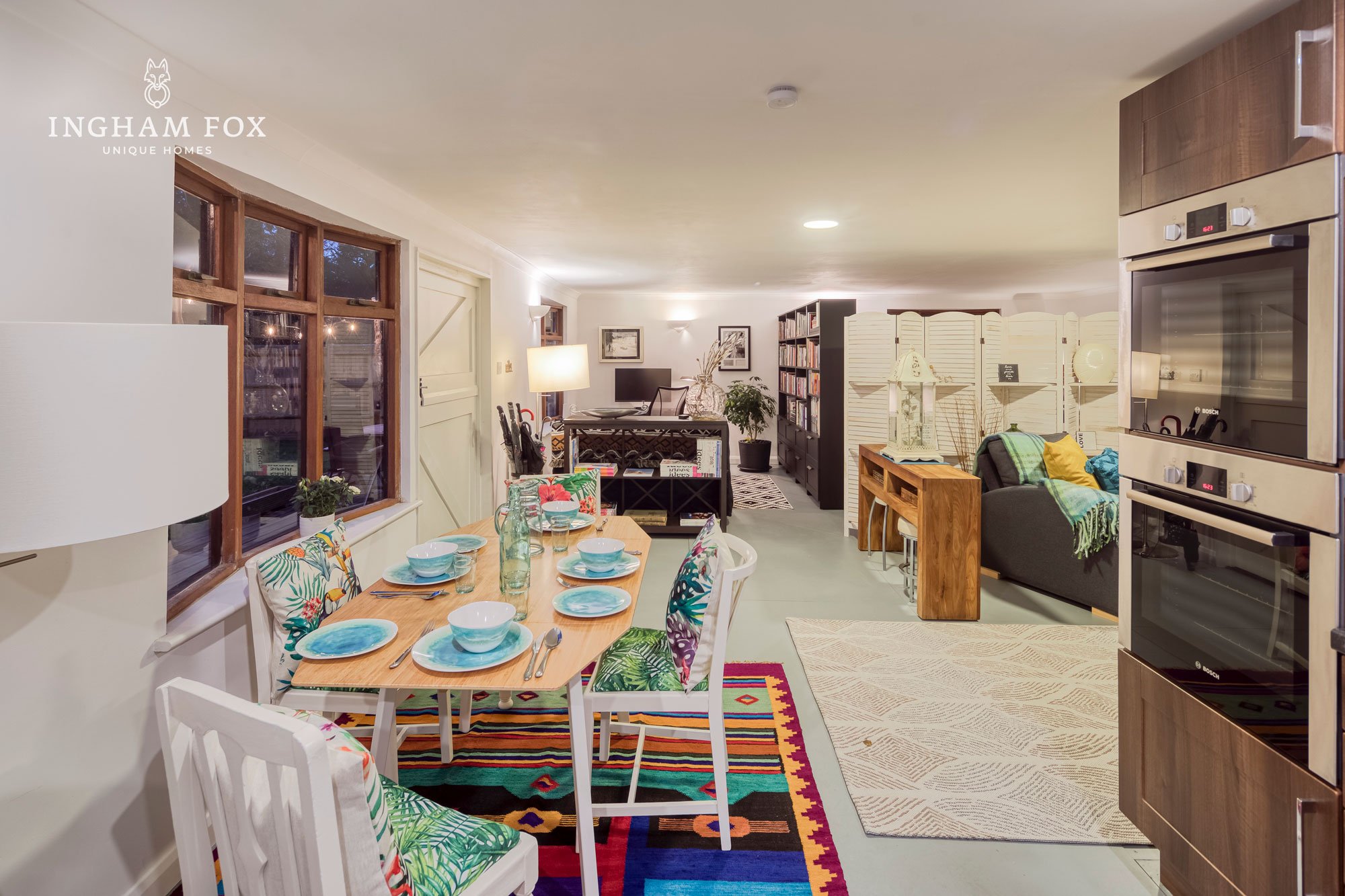 Modern open-plan kitchen and living area with colorful dining setup, wooden table, vibrant cushions, and decorative accents.