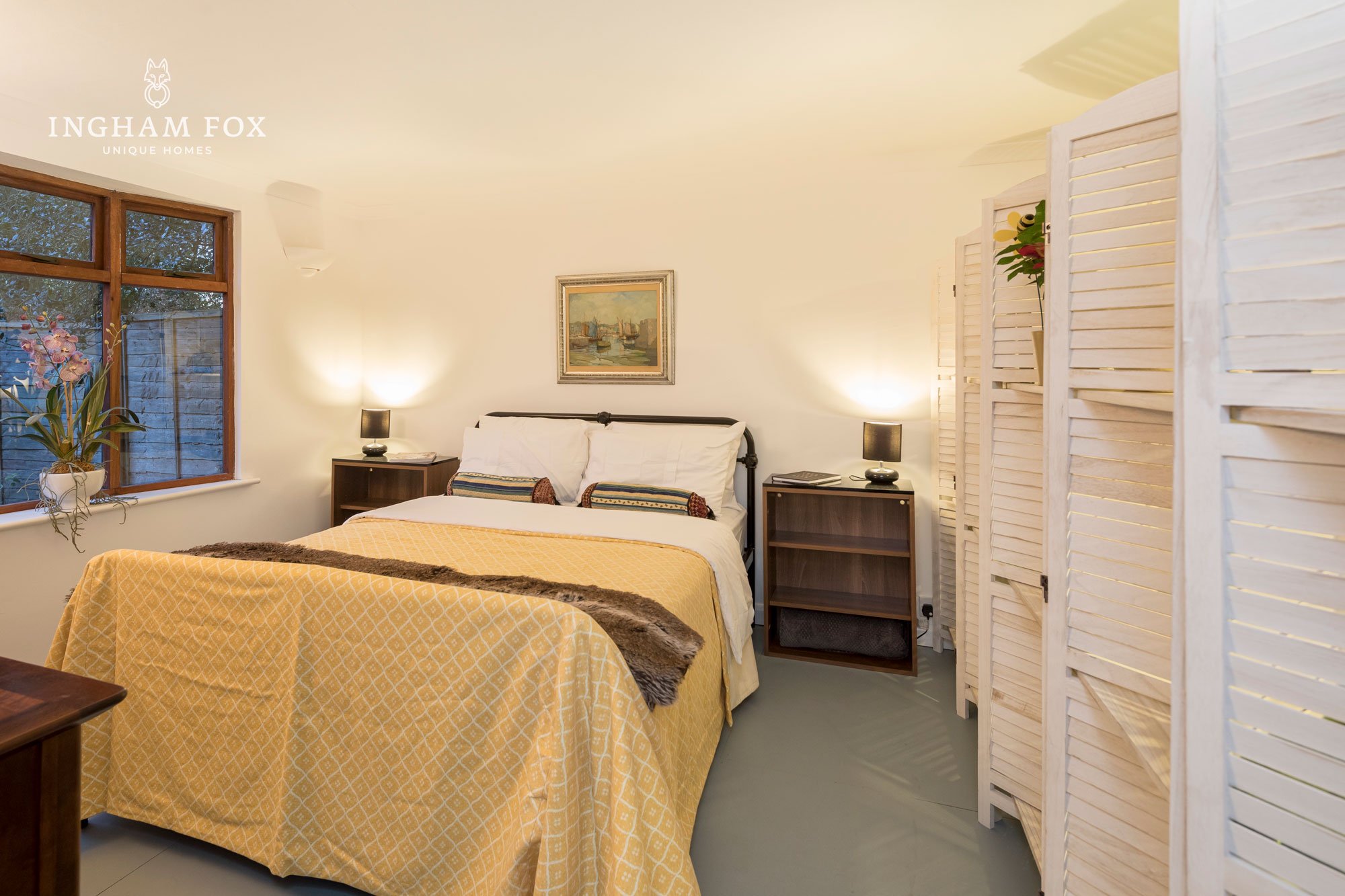 Cozy bedroom with a bed, yellow bedspread, bedside tables, lamps, painting on wall, window with flowers, and room divider.