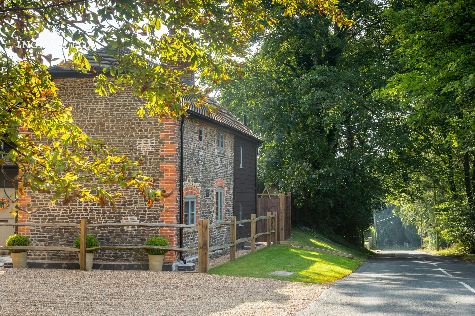 A rustic brick house with wooden accents, surrounded by trees and greenery, near a quiet road. Sunlight filters through the leaves, casting shadows on the ground.